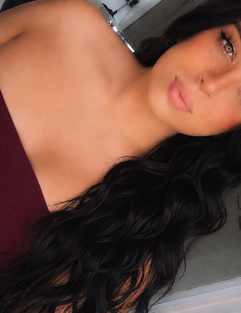 A woman with long curly black hair and green eyes, showing her face and bare shoulders, wearing a maroon top.