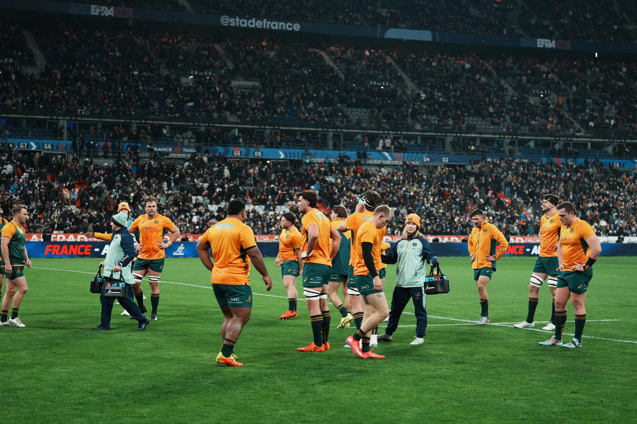 Australian rugby team on the field during a match at Stade France, with fans in the stadium stands in the background. Eliza Freney, sports dietitian working in elite rugby union is handing out hydration to Taniela Tupou, Harry Wilson and team members