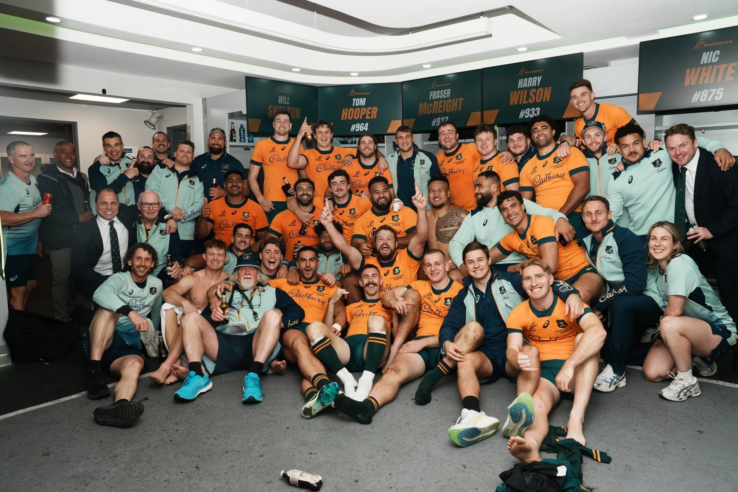 A group of Australian rugby players and staff members celebrating in the change rooms after a historic win in Johannesburg against the South African team, the 'Boks' in 2025.