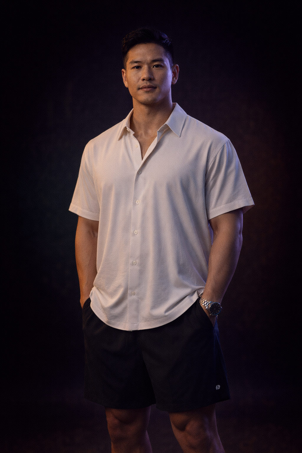 A young man standing confidently with hands in his pockets, dressed in a white short-sleeve button-up shirt and black shorts, wearing a watch, against a dark background with subtle lighting.