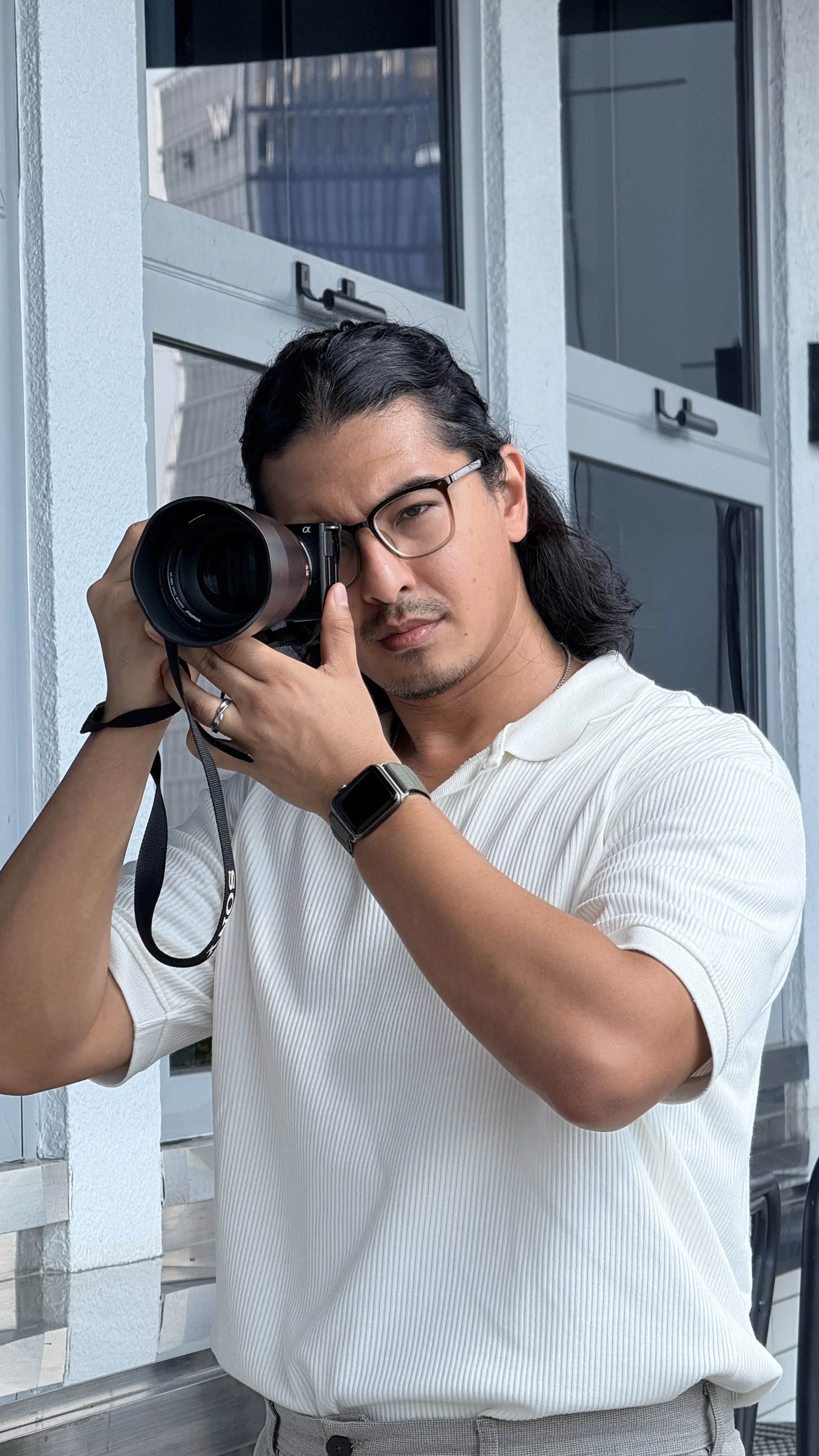Andrew is holding a camera up to his eye, standing outdoors near a building with large windows.