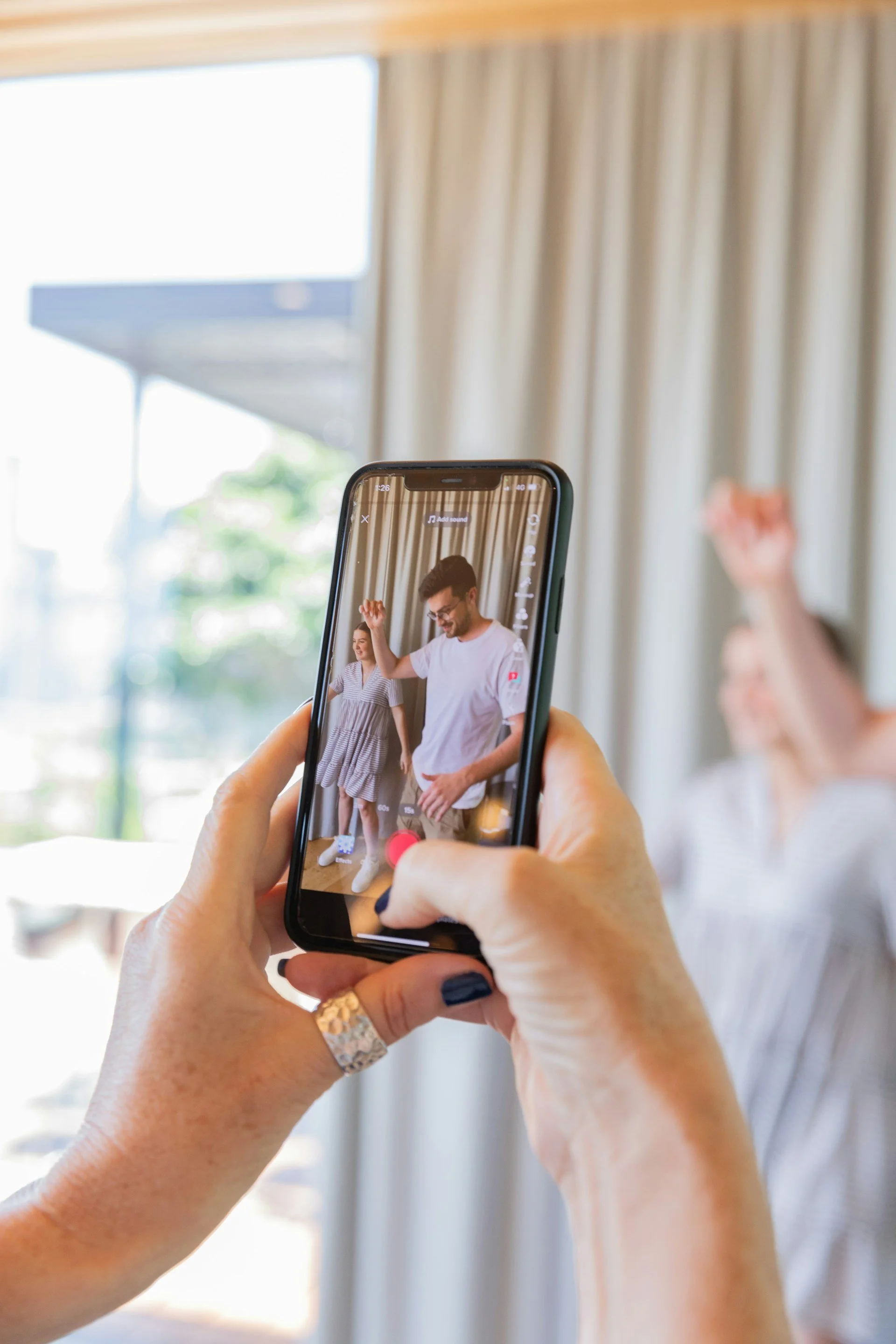 Person taking a photo of a man and woman dancing indoors, with large windows and curtains in the background.