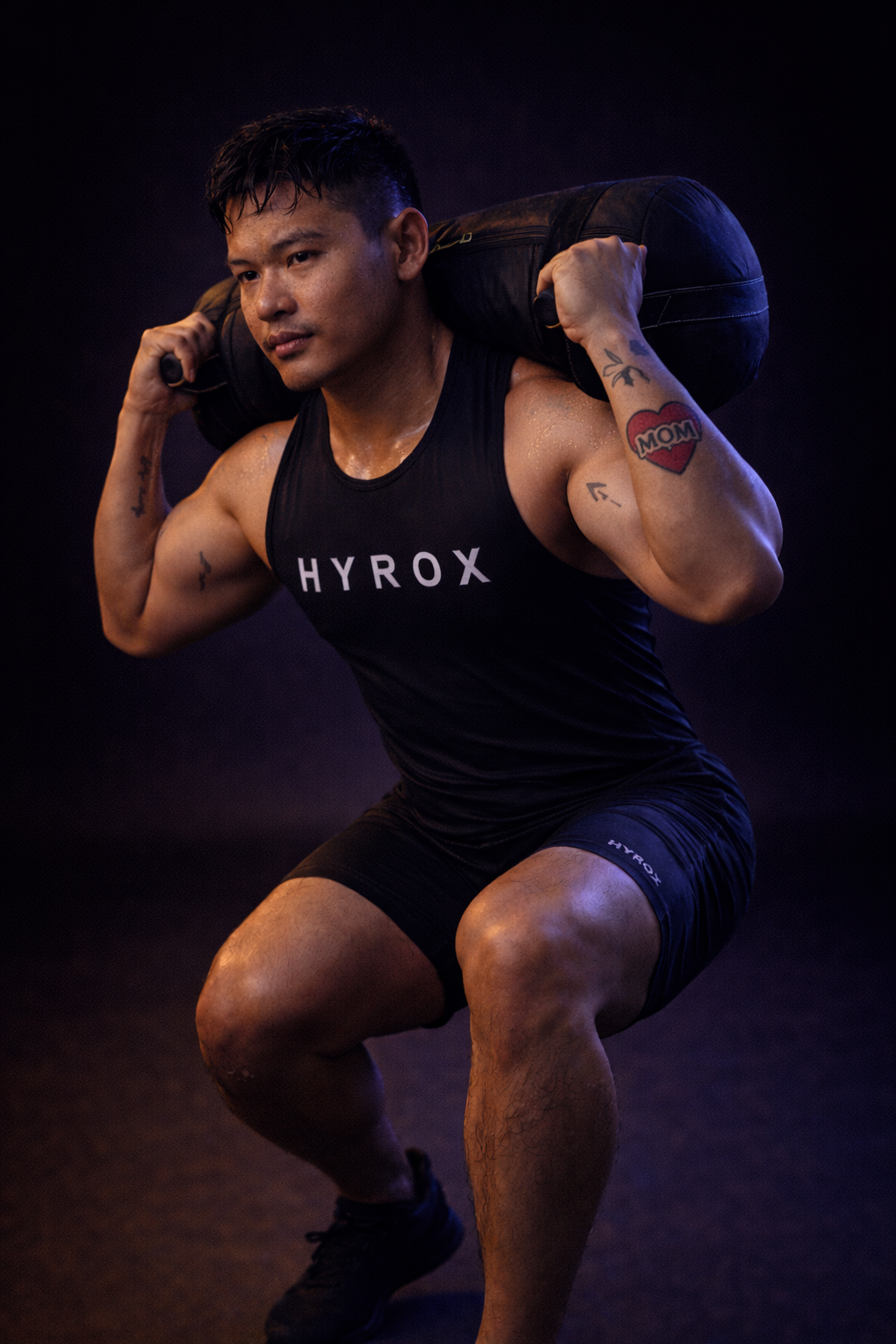 A muscular man with tattoos, wearing a black tank top and shorts, carrying a large black sandbag on his shoulders during a workout in a gym.
