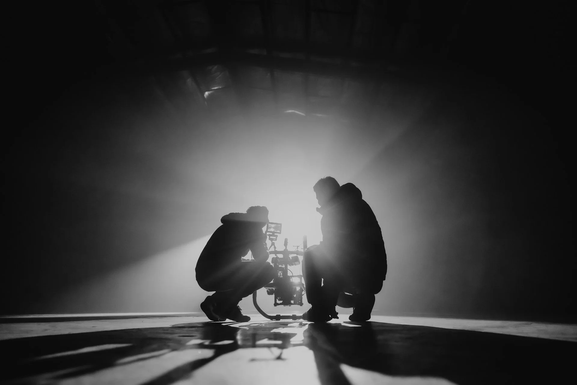 Two people working with a camera on a tripod in a dimly lit indoor space, backlit creating silhouette effects.