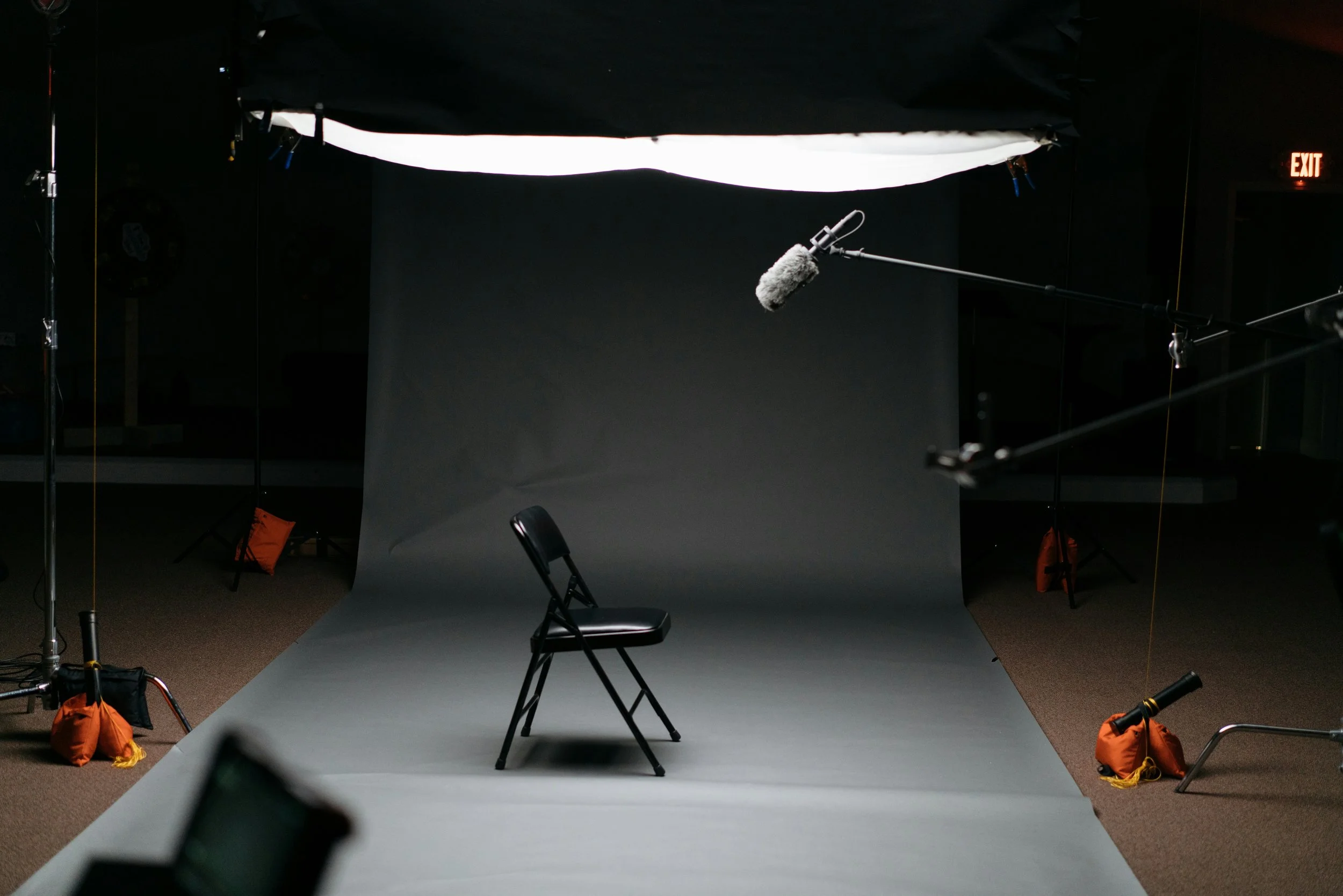 An empty black chair on a photography studio grey backdrop with a boom microphone and studio lighting equipment.