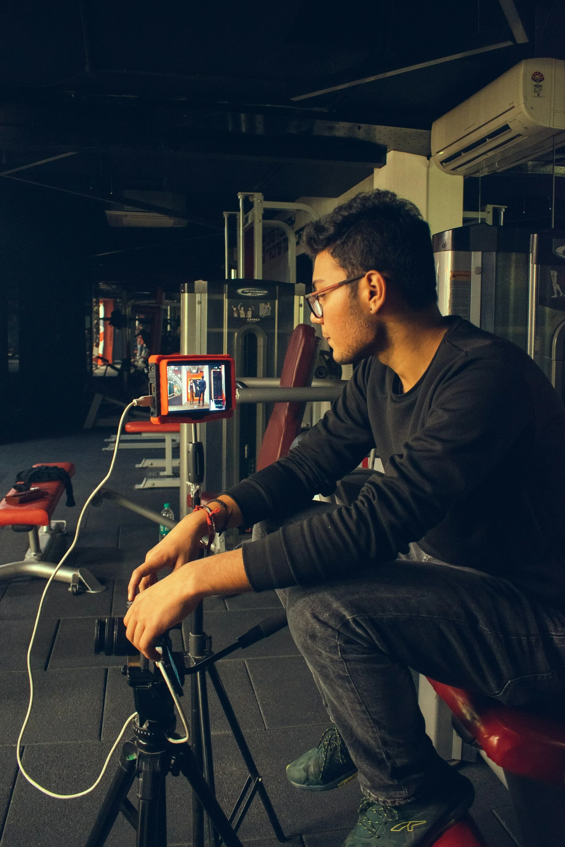 A young man with glasses and dark hair sitting in a gym, filming himself with a camera mounted on a tripod, with gym equipment in the background.