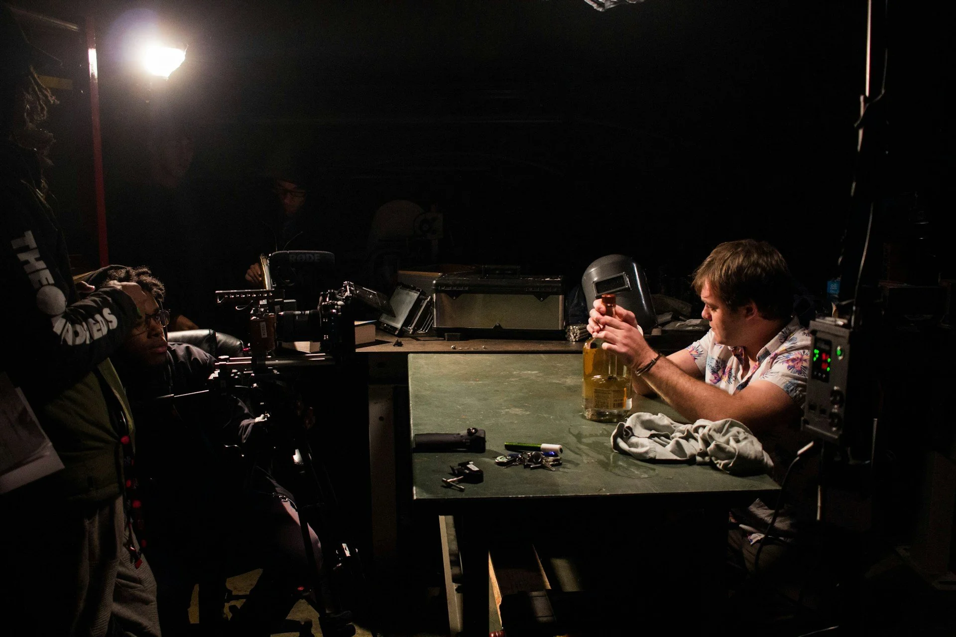 A man with light brown hair sitting at a green table in a dark room, holding a bottle of alcohol. Several small items like keys and a marker are on the table. Another person is operating a camera or filming equipment, with accompanying technical gear around. The scene appears to be backstage or in a studio setting.