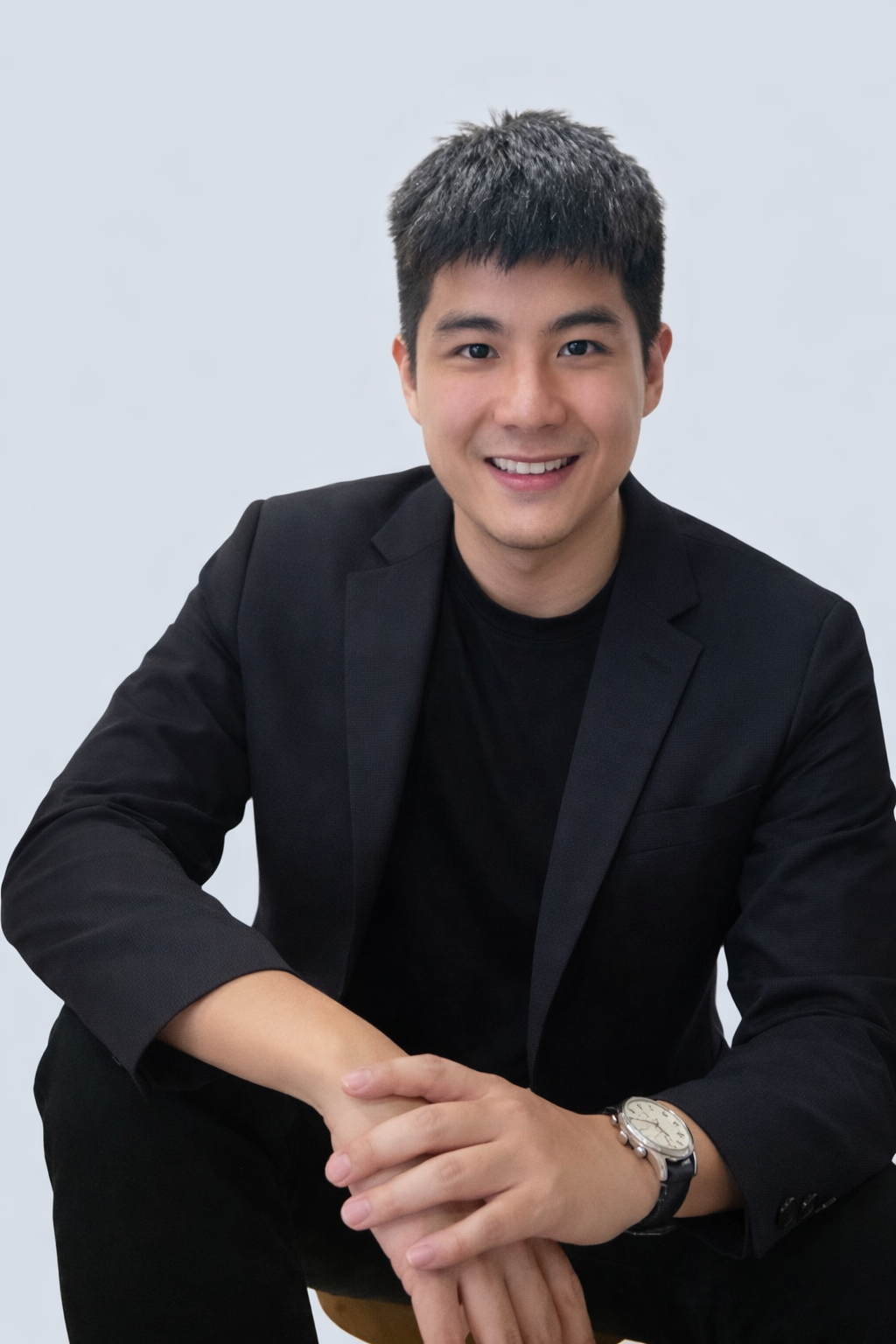 Nicholas in black blazer and watch smiling, seated against a plain light gray background.