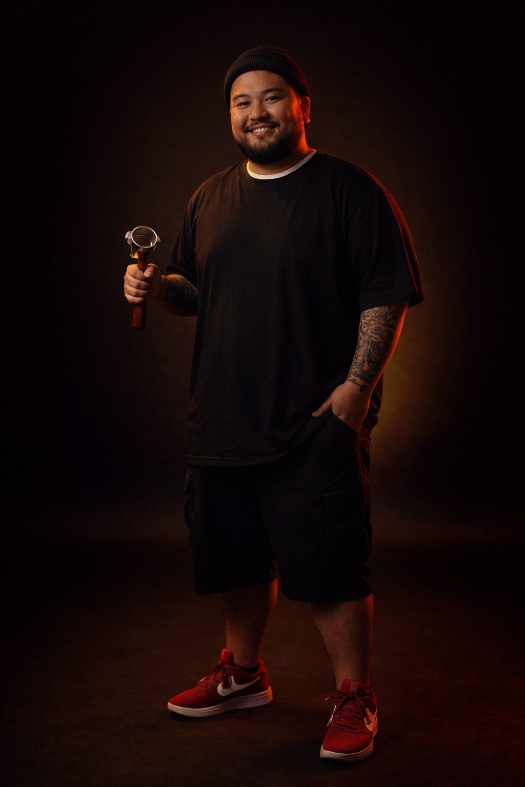 A man wearing a black beanie, black T-shirt, and black shorts, holding a hammer in his right hand, standing against a dark background with a warm glow.