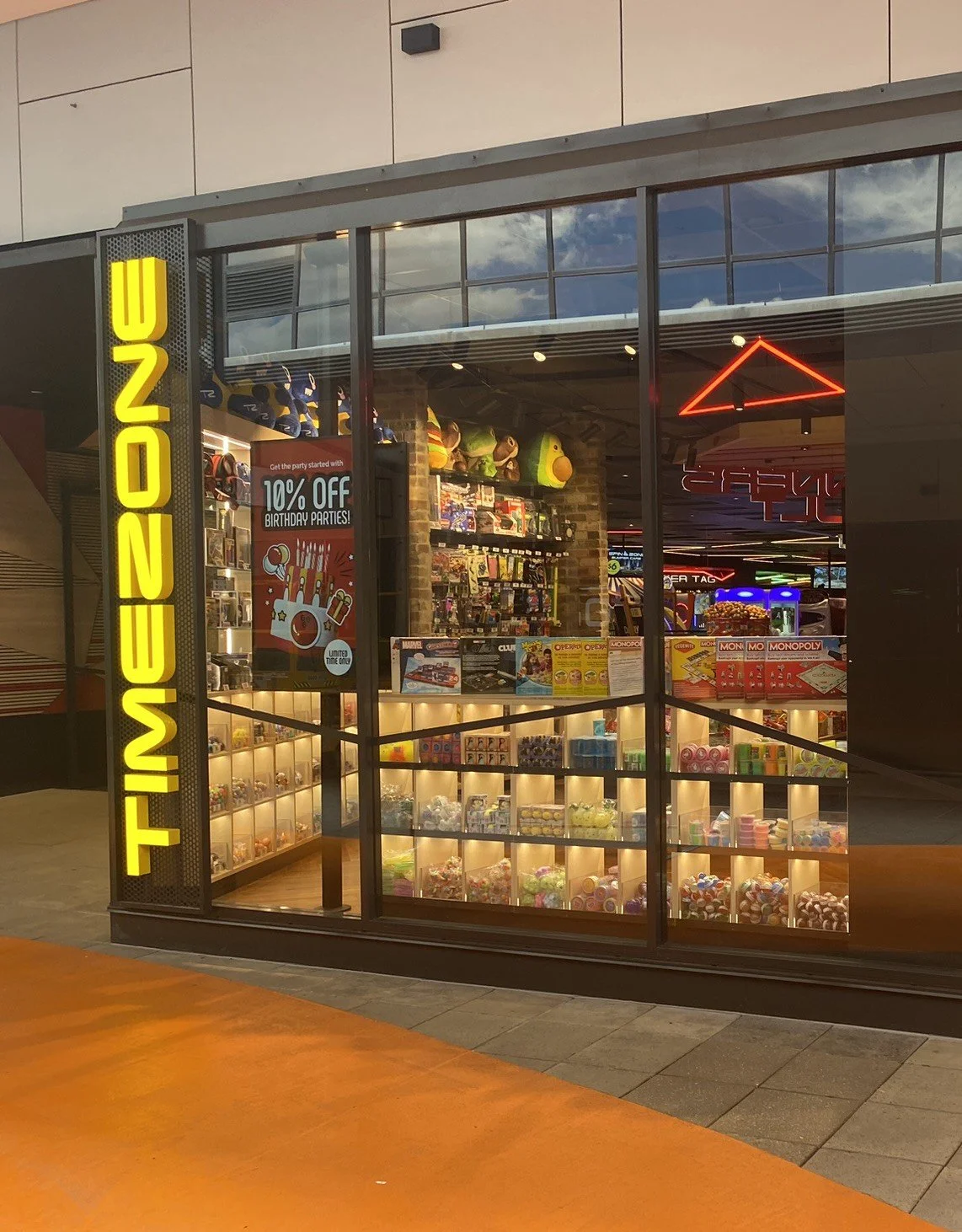 Toy store with a bright yellow vertical sign saying 'TINY TOWN'. Inside, shelves display various toys including puzzles, plush toys, and board games. Promotional sign offers 10% off birthday parties. The store has glass windows, neon lights, and a wooden floor at the entrance.