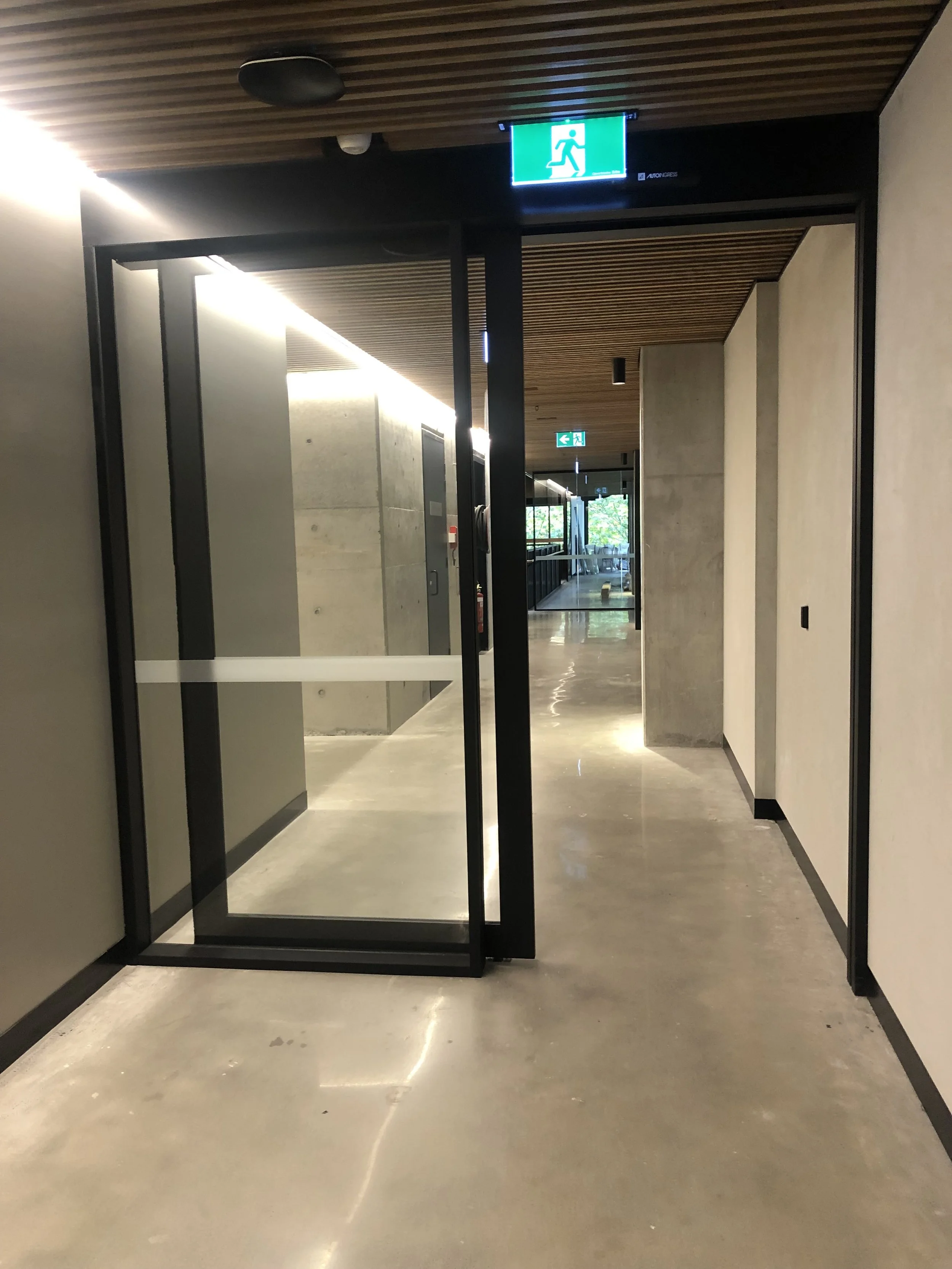 An indoor scene with a glass door and a green emergency exit sign above it, leading to a corridor with natural light coming through glass doors at the end.