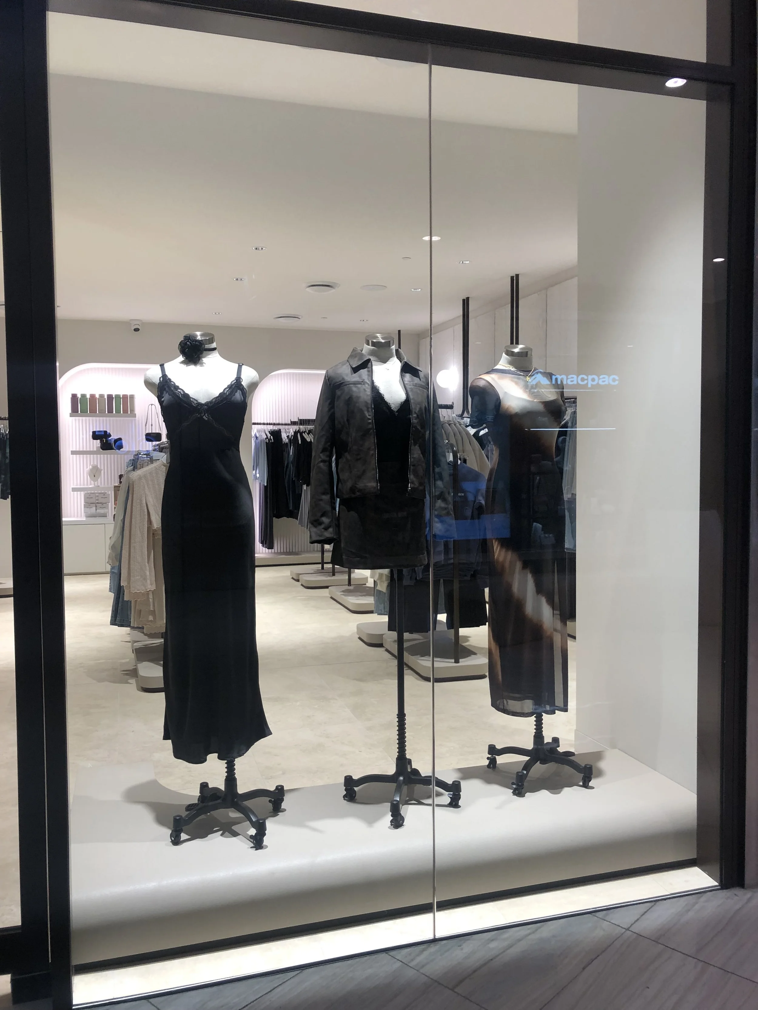 Display window with three mannequins dressed in black outfits inside a clothing store. The background shows shelves of colorful items and hanging racks of clothes.