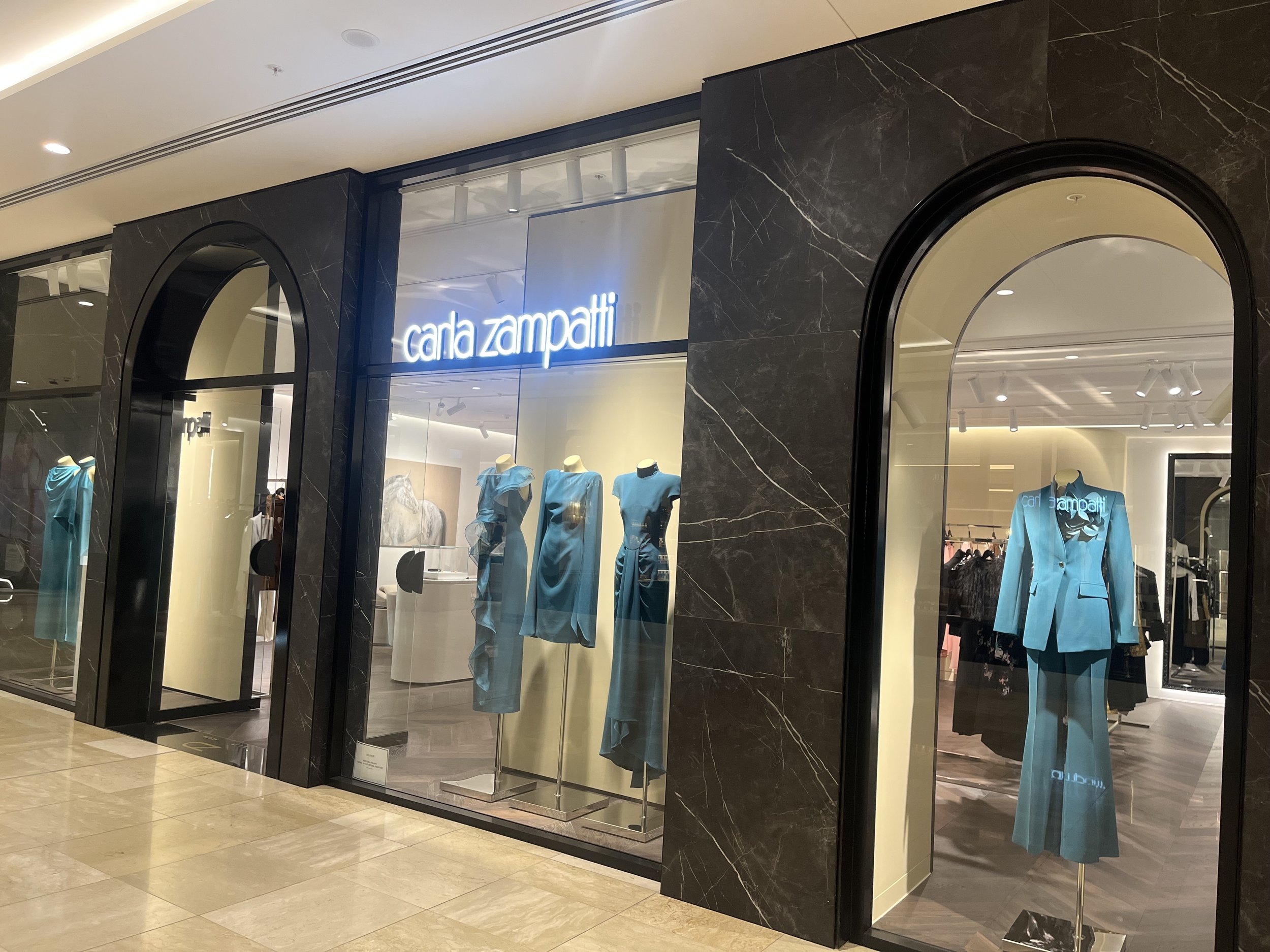 Front view of Carla Zampatti boutique with mannequins in blue outfits on display inside, large glass windows, and black marble accents.