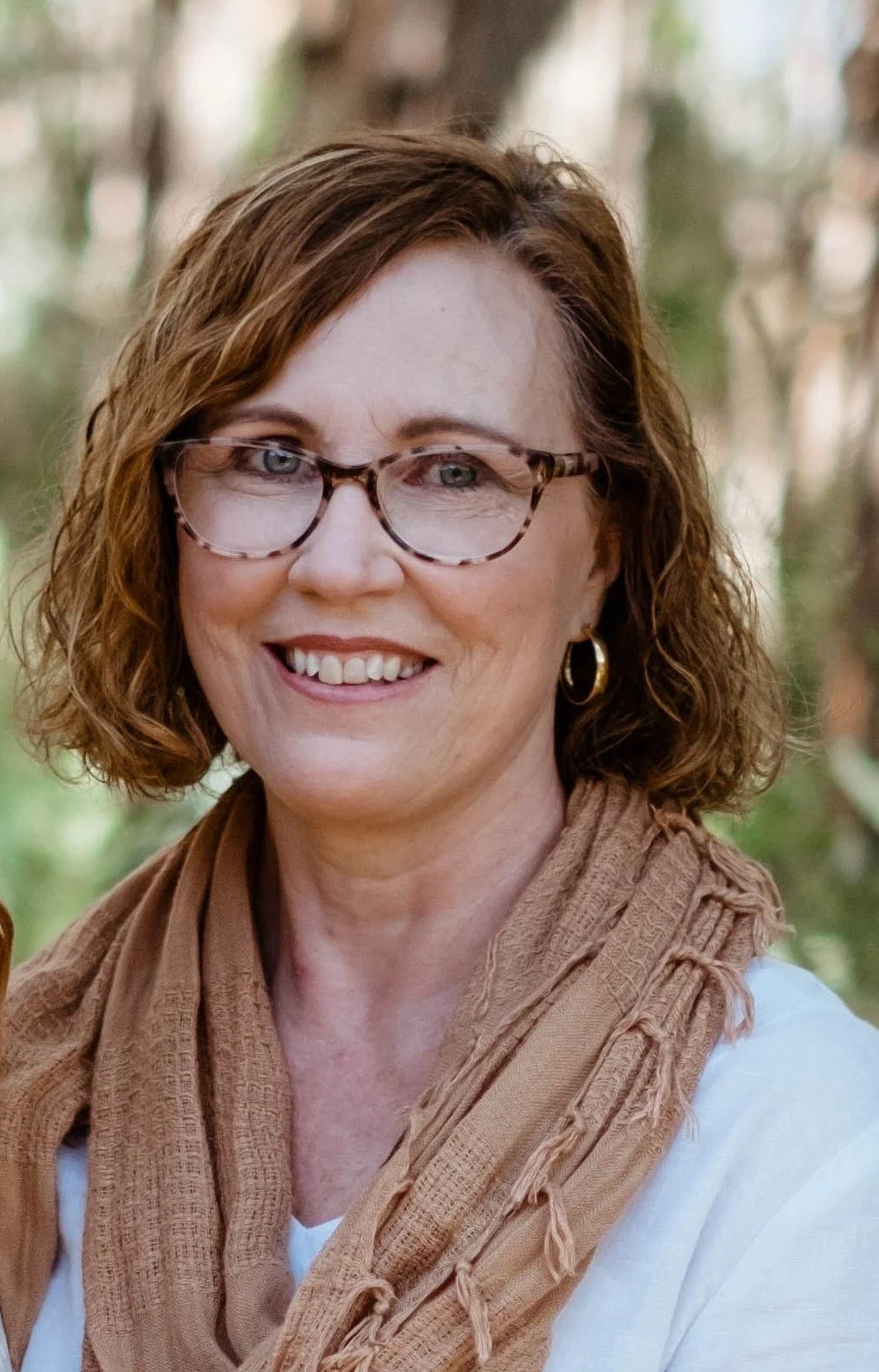 A woman with curly brown hair, wearing glasses and gold hoop earrings, smiling outdoors with a blurred background of trees.