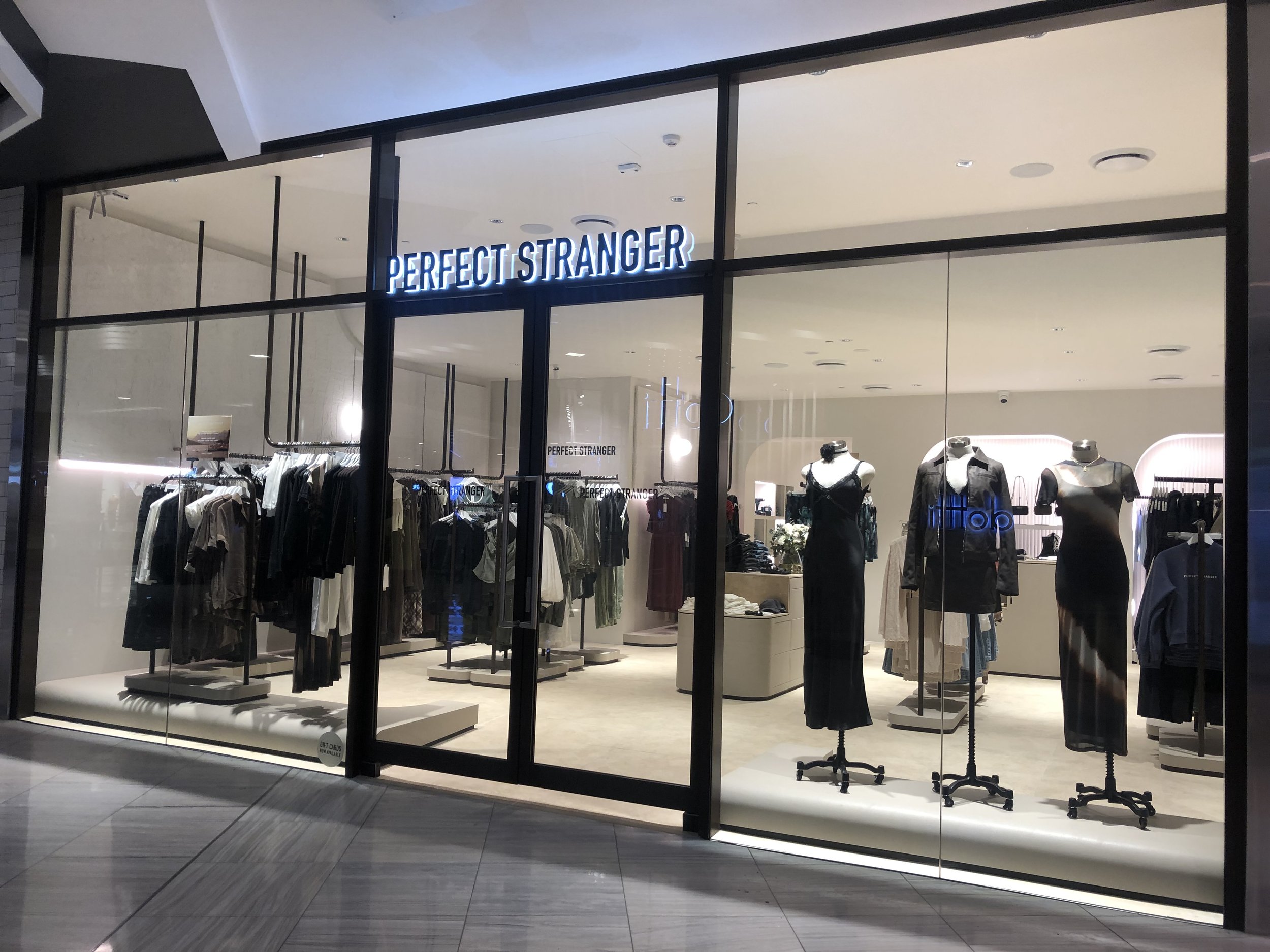 Clothing store front named 'Perfect Stranger' with mannequins dressed in black dresses, racks of clothes, and bright retail lighting inside.