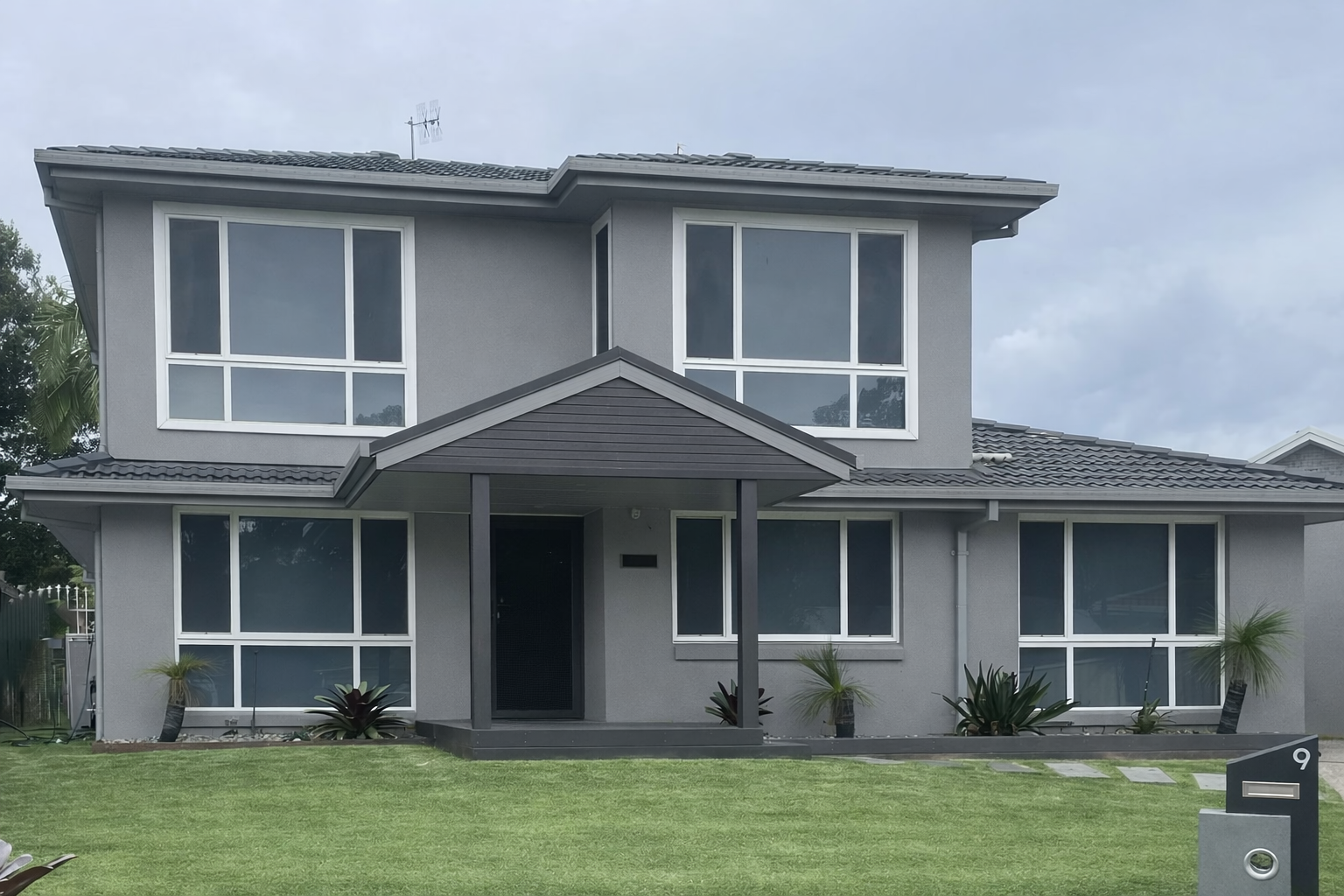 Front view of a modern two-story gray house with large windows, a small front porch, and a well-maintained green lawn with plants and a mailbox in the yard.