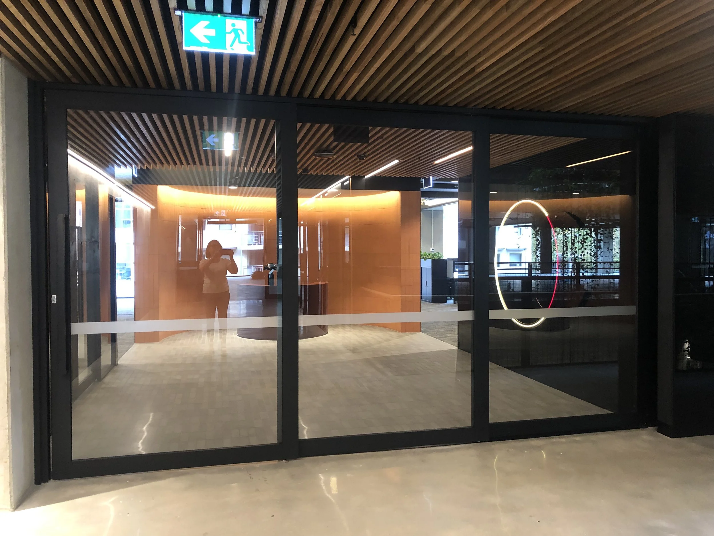 Glass entrance doors to a modern building lobby with a blue exit sign above, showing a person running towards an emergency exit.