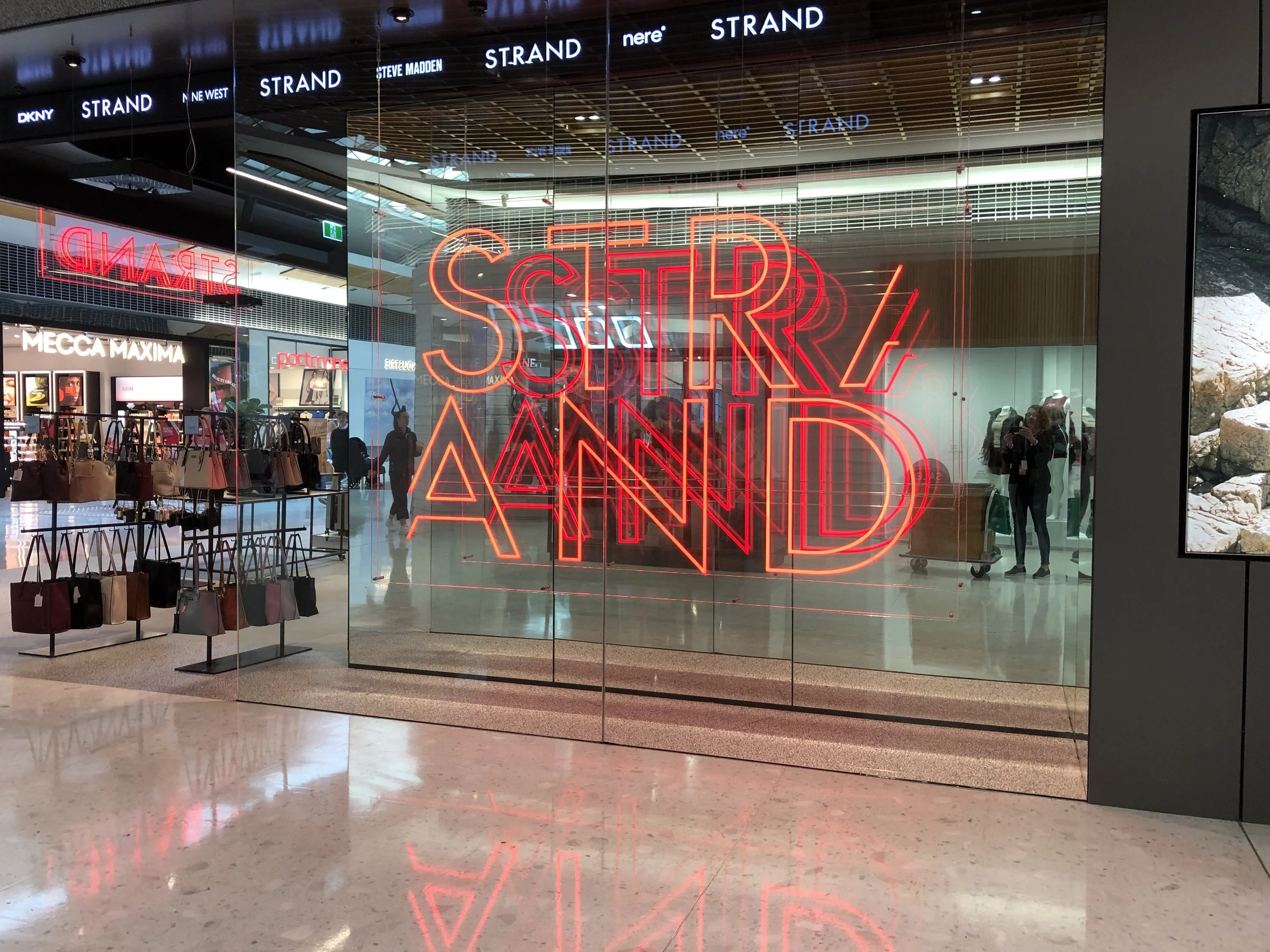 Neon sign in a shopping mall window that reads 'STIR AND' with reflections on the glass and stores visible inside.