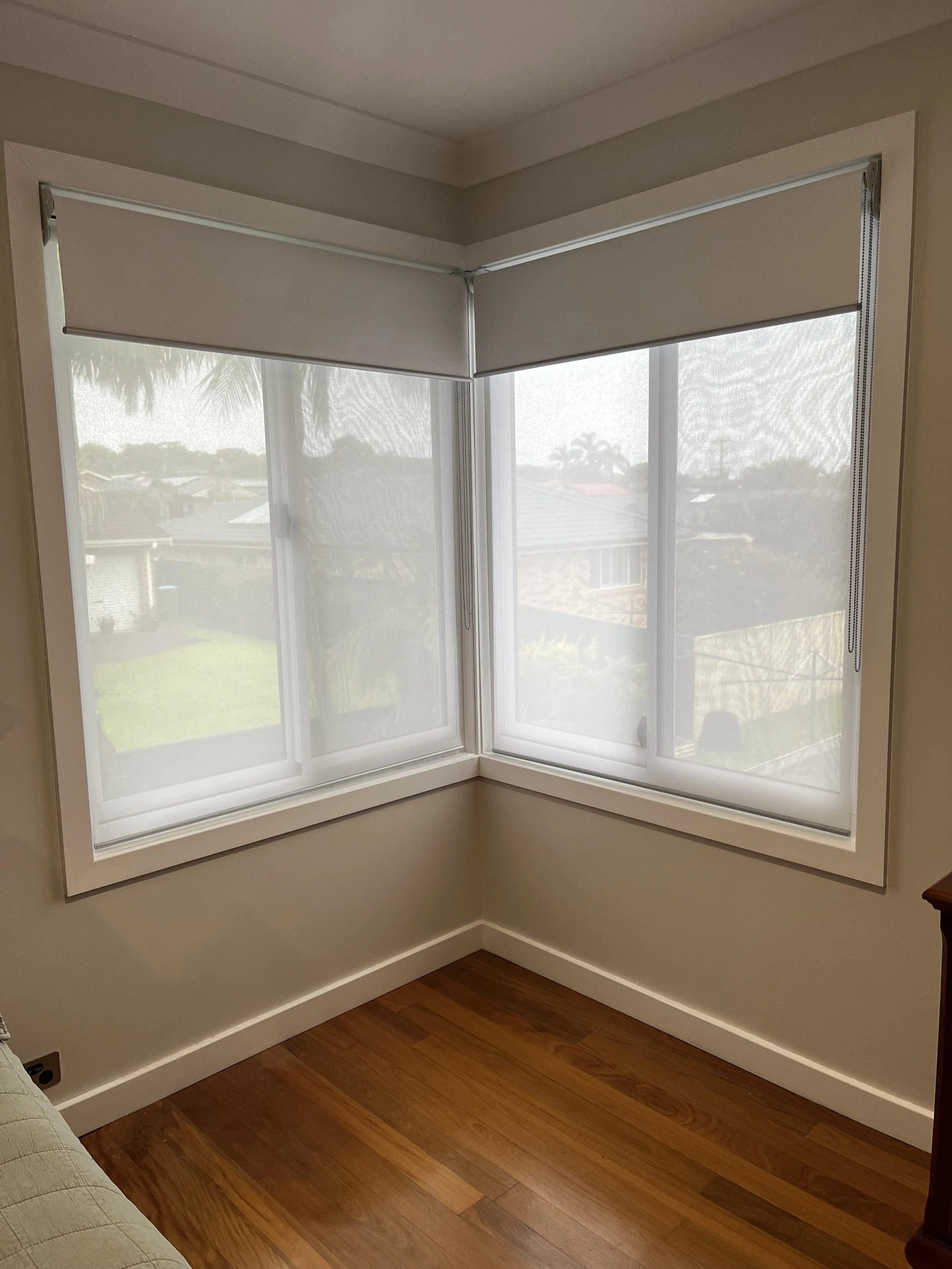 A room with a large bay window covered with white roller shades, showing a view of a backyard with grass and neighboring houses.