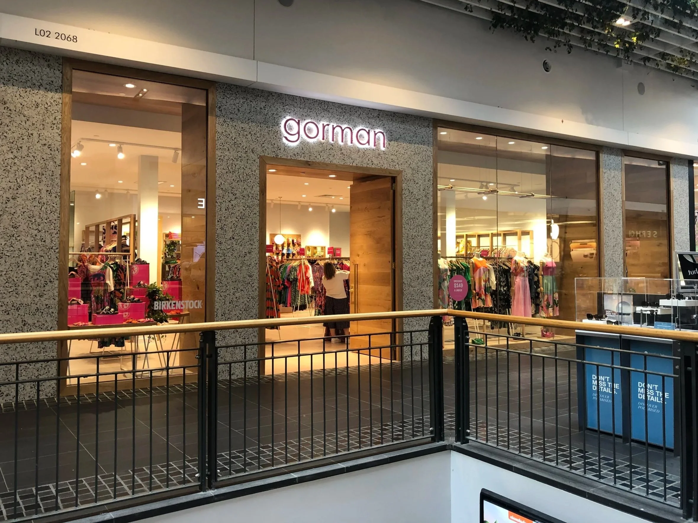 The storefront of a clothing store called Gorman, with large glass windows displaying racks of brightly colored clothing inside. The store's sign is illuminated, and the entrance is open. There is a black railing in front of the shop and a blue recycling bin nearby.