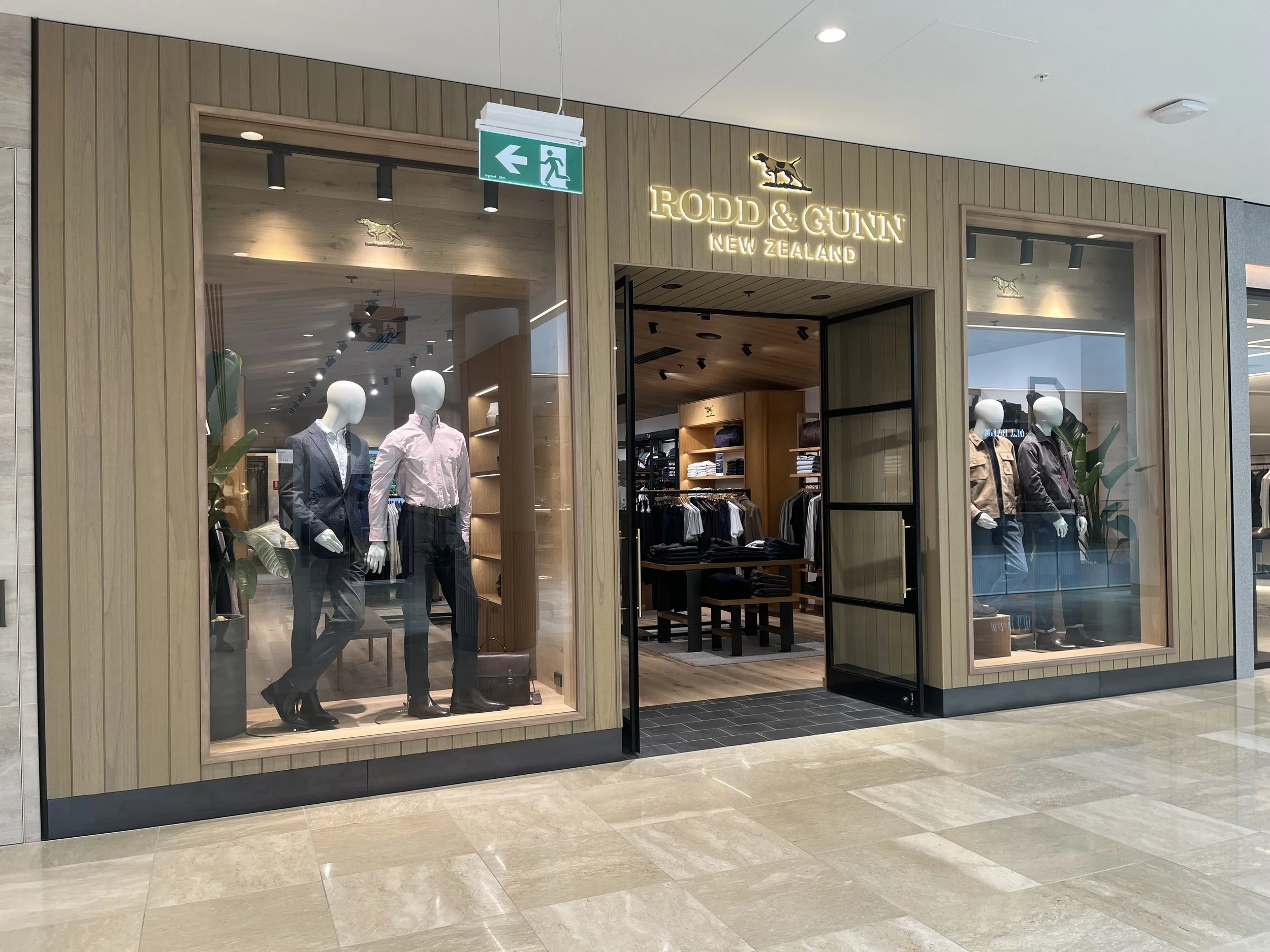 Storefront of Rodd & Gunn clothing store with mannequins in the window displaying men's suits and casual wear, wood-paneled exterior, neon sign with the store's name, and an emergency exit sign above the entrance.