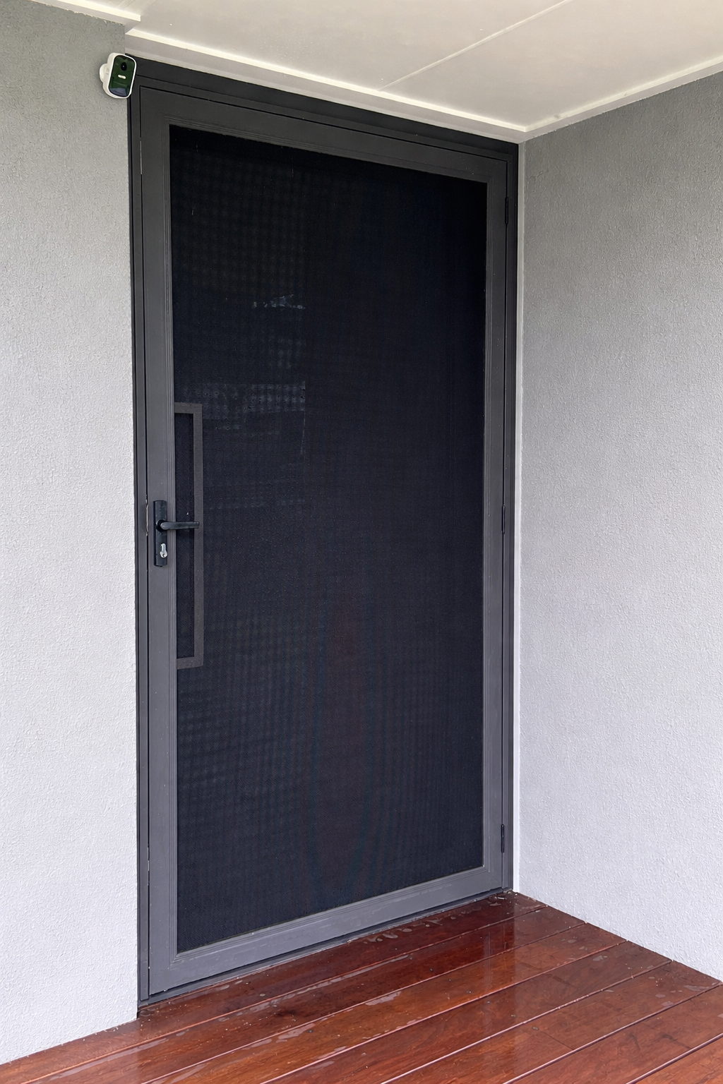 A black screen door with a screen panel, installed in a gray wall, with a wooden deck at the bottom and a small animated security camera in the top left corner.