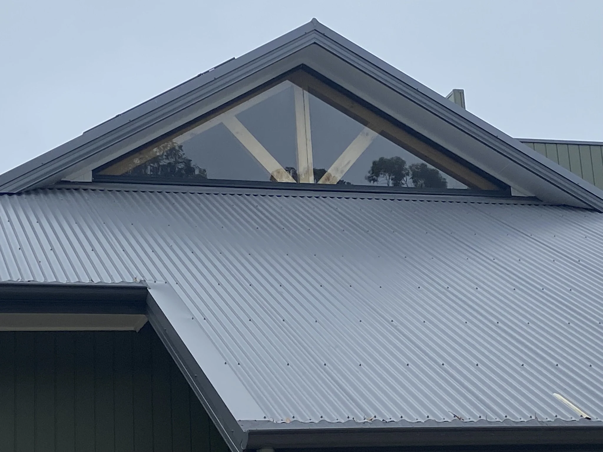 Close-up of a modern house roof with metal siding and a triangular window at the peak, showing trees reflected in the glass.