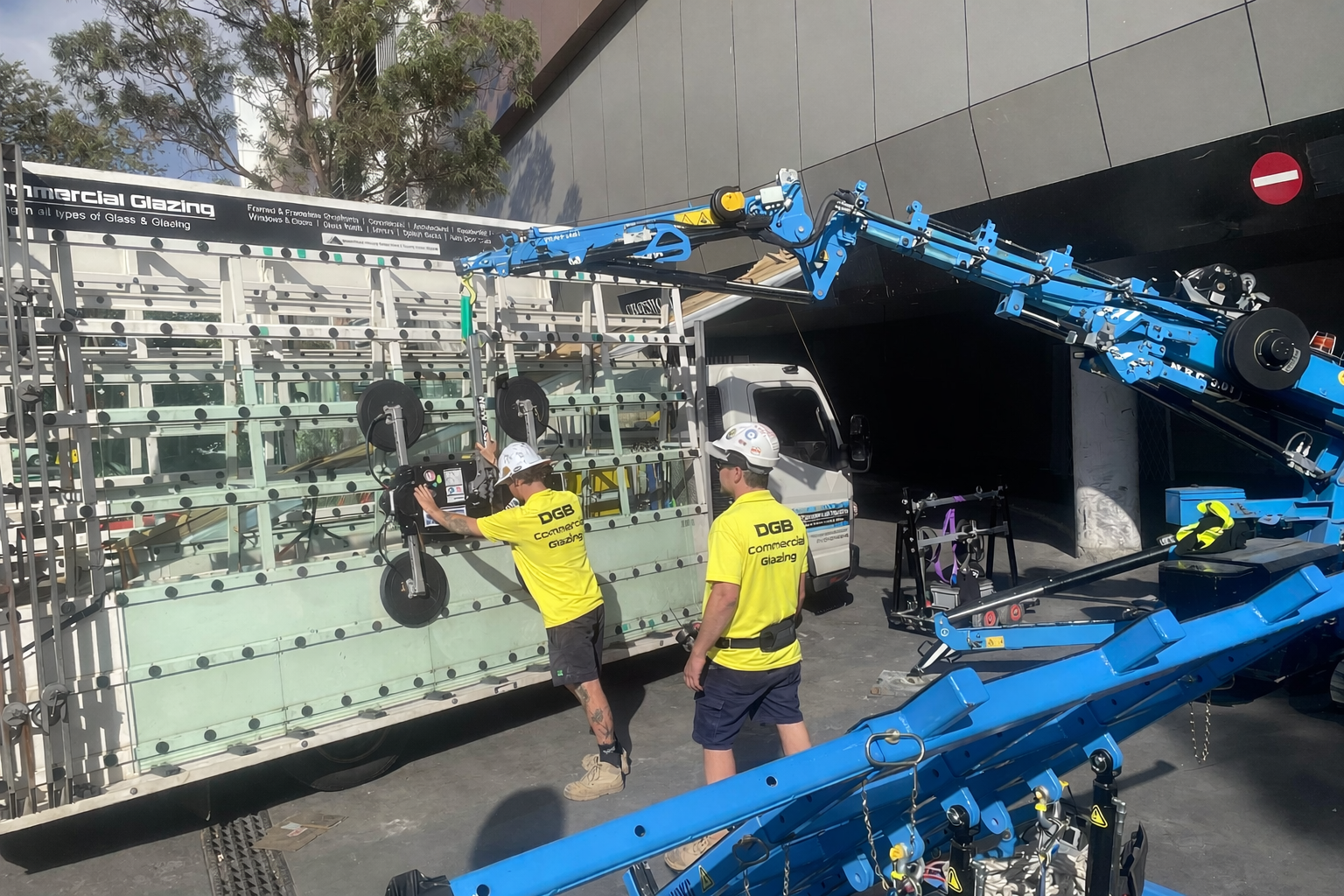 Two workers in yellow shirts and helmets installing or repairing glass panels on a large glass structure, with a blue crane truck assisting in the process, near a building entrance.