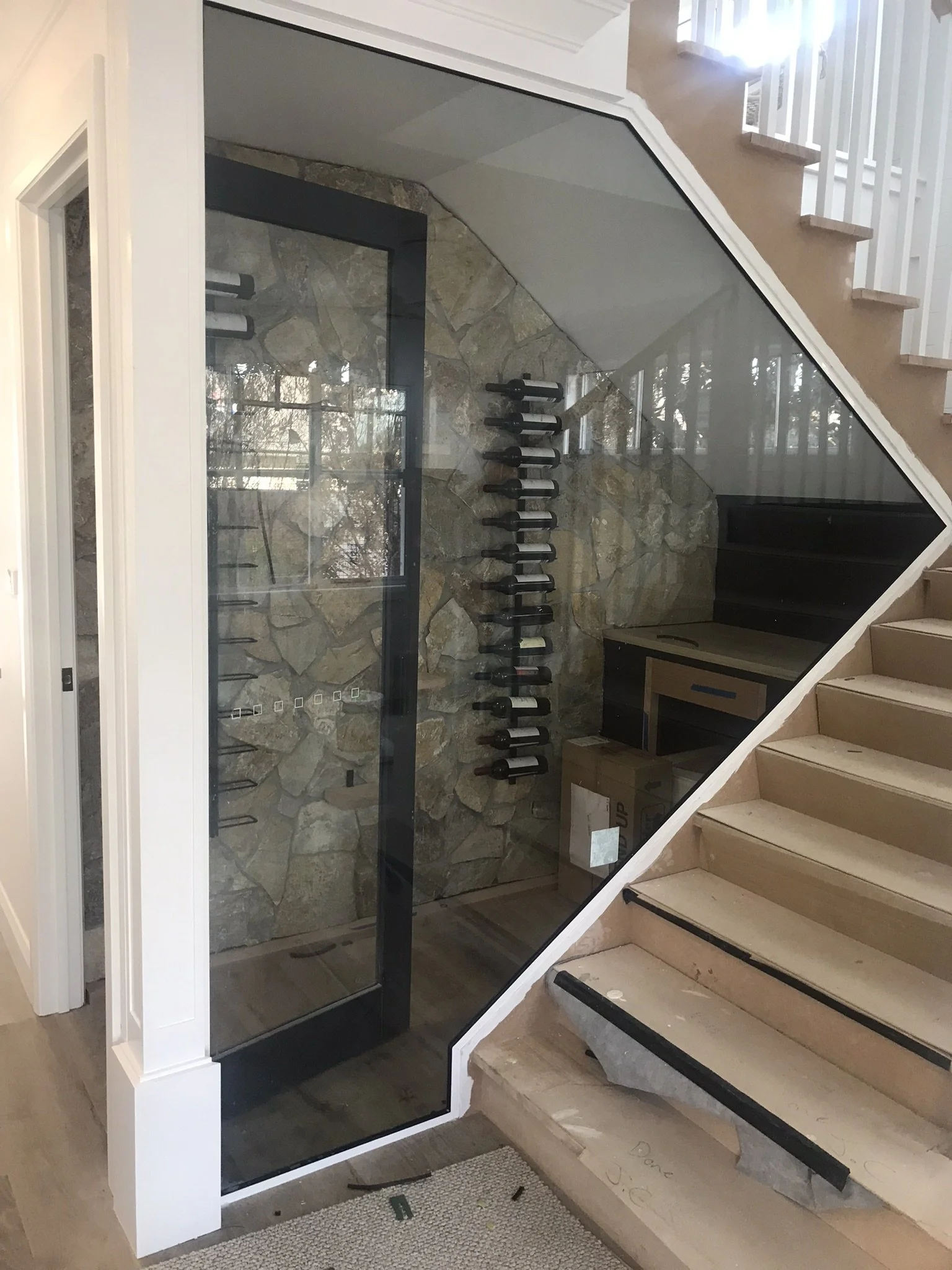 Interior view of a staircase with wooden steps and a glass railing, featuring a wine rack on a stone wall underneath the stairs and a small desk or counter area in the corner.