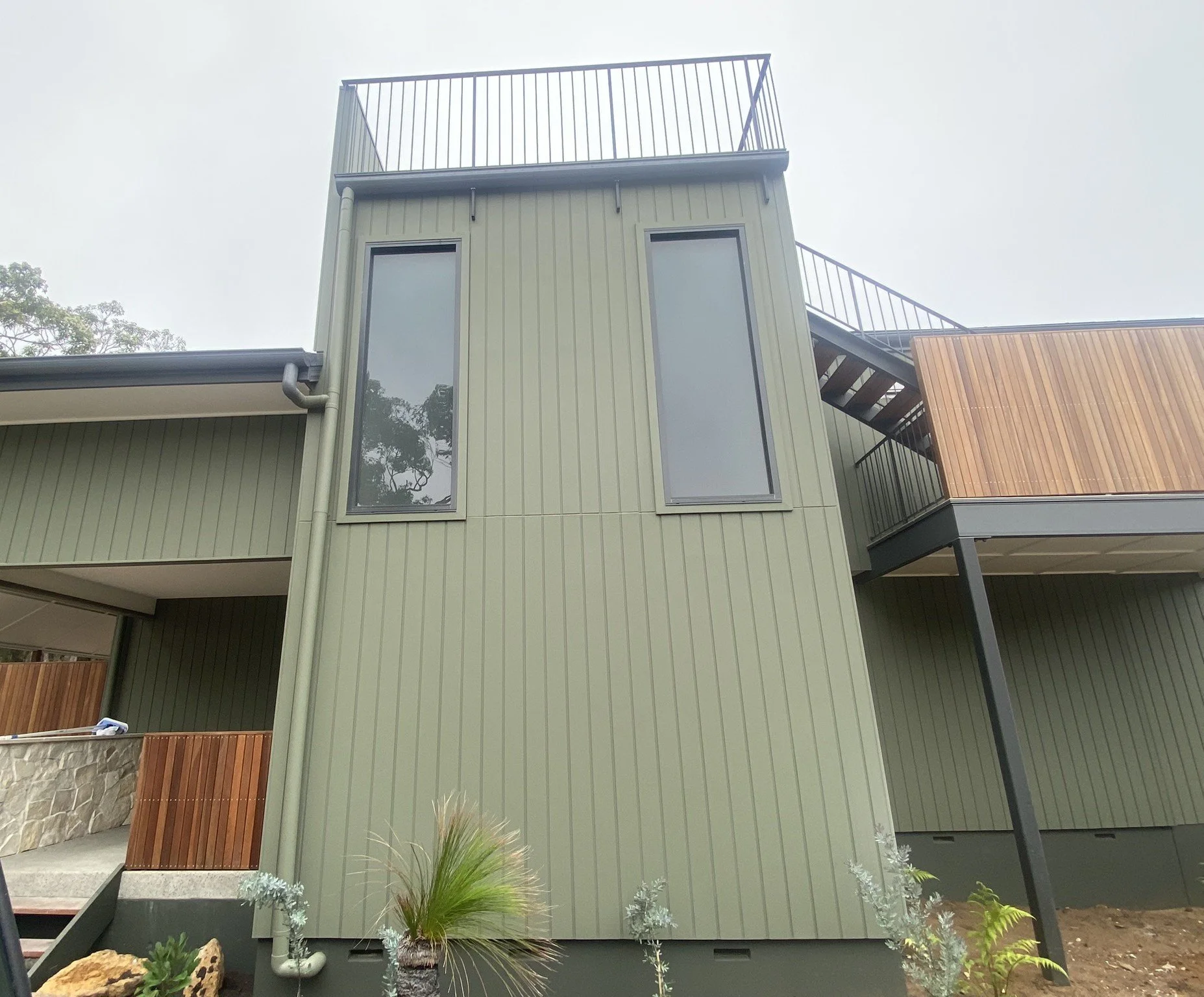 Modern multi-level house with green siding, large windows, and a rooftop balcony, with black railings and a staircase.