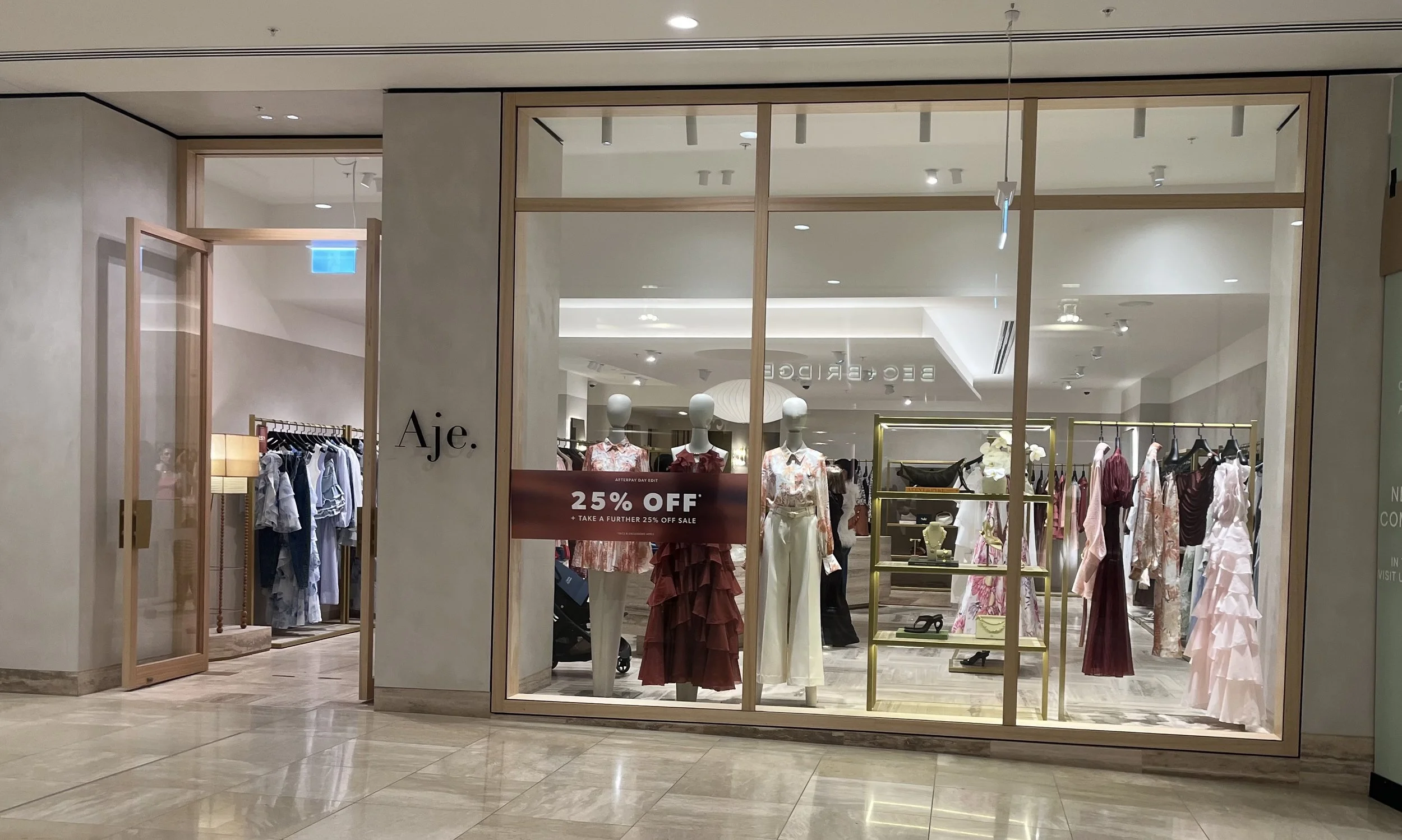 Clothing store display window with mannequins wearing dresses, sale sign offering 25% off, and racks of clothes inside the store.