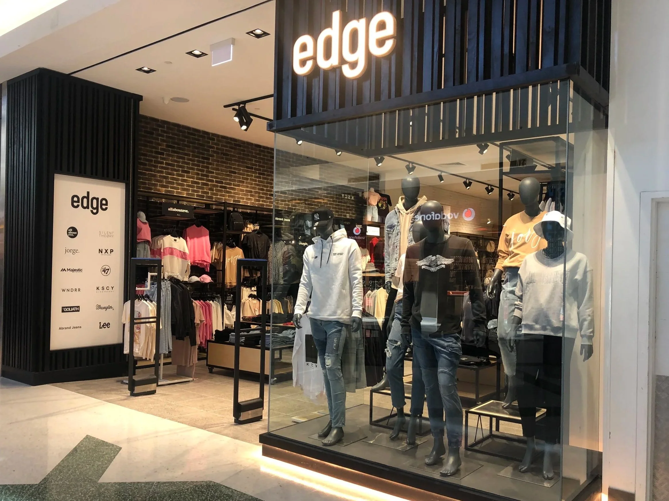Storefront display window of Edge clothing store with mannequins dressed in casual wear, and a sign listing brands like All About Eve, Jorge, NXP, and Lee.