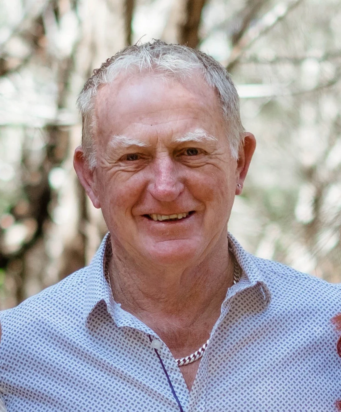 Close-up of an older man smiling outdoors, wearing a light patterned shirt and a silver chain necklace, with a blurred natural background.