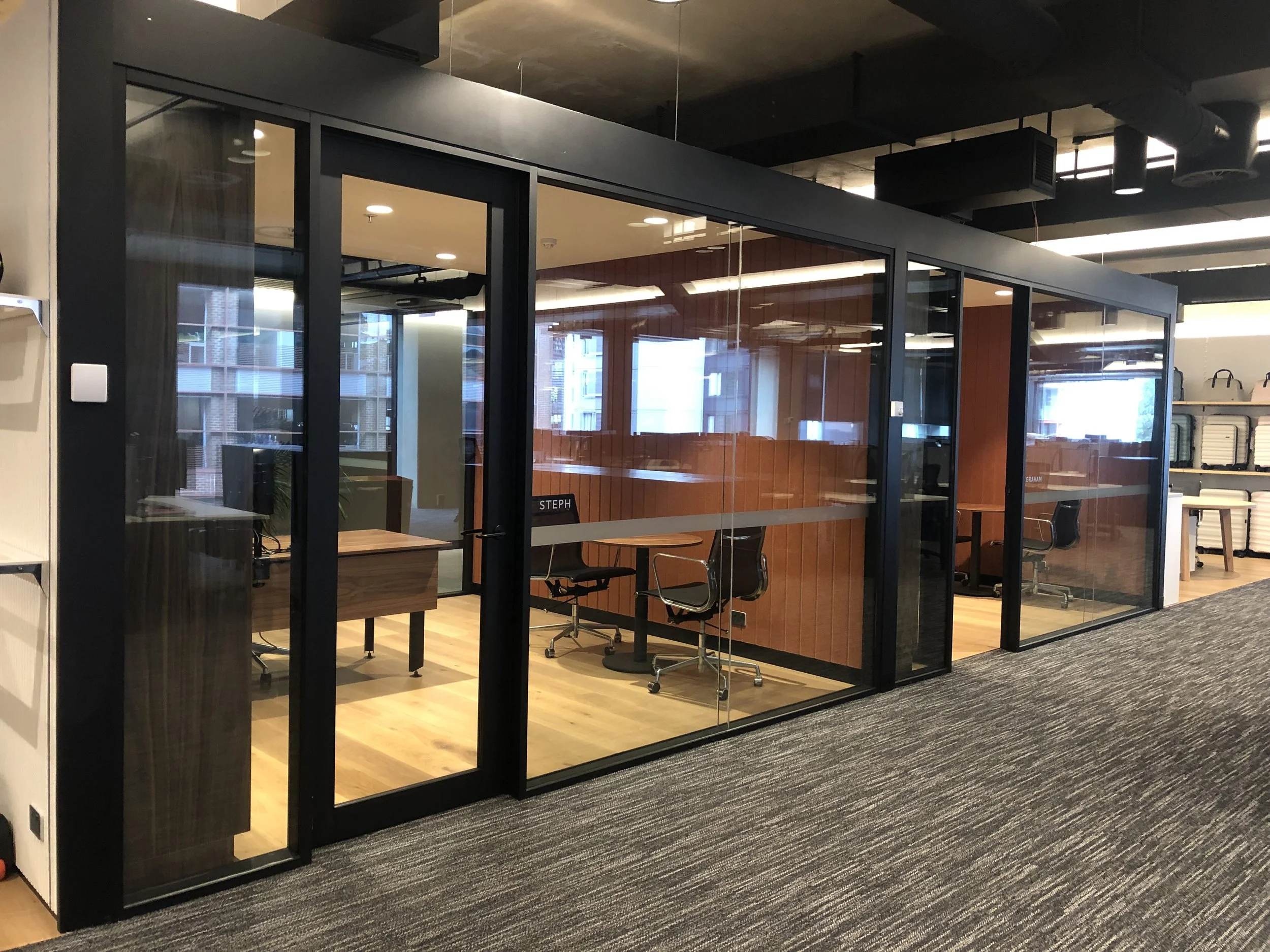 An office space with glass-walled conference rooms, containing desks and chairs, situated on a wooden floor near a carpeted area.