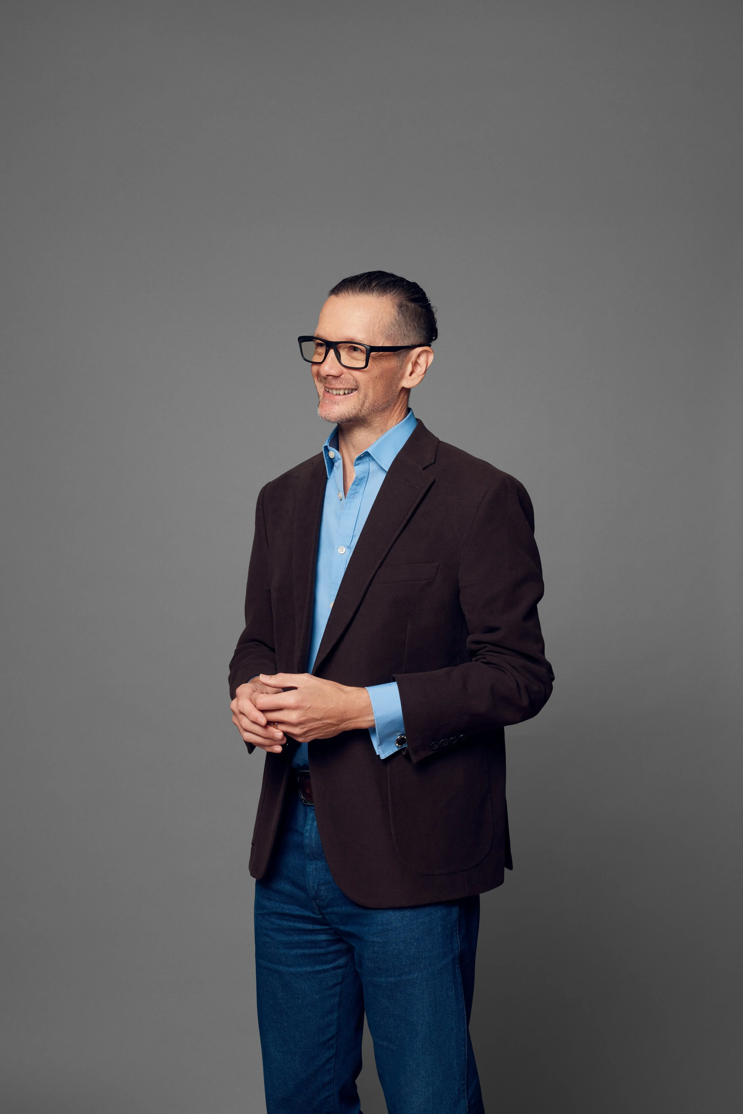 A smiling man with dark hair styled in a slicked-back fashion, wearing glasses, a dark blazer, a light blue button-up shirt, and blue jeans, standing against a gray background.