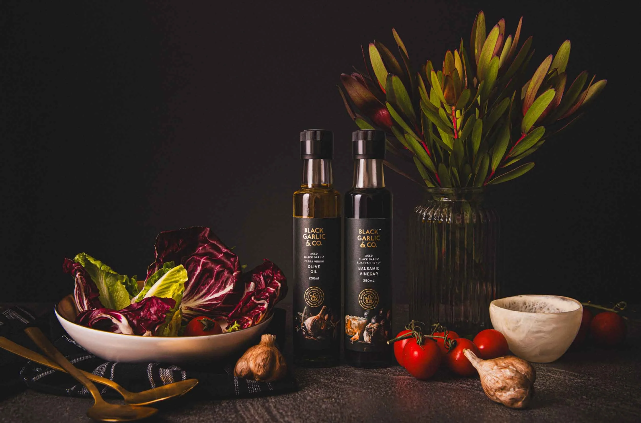 A stylised photograph of Black Garlic & Co's Olive Oil and Balsamic Vinegar bottles pictured with black garlic, vine ripened tomatoes and lettuce leaves in a large serving bowl