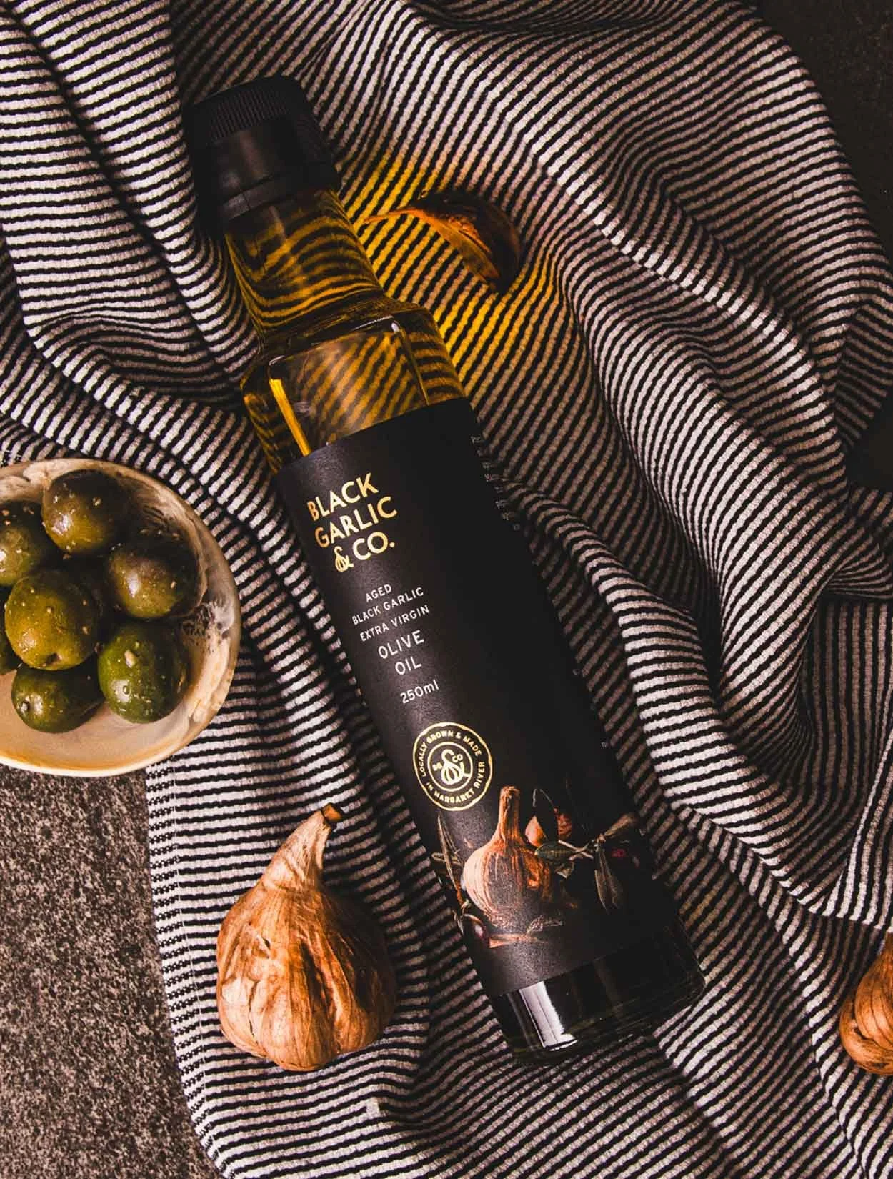 A flat lay stylised photograph of Black Garlic & Co's Aged Black Garlic Extra Virgin Olive oil bottle, showcasing the brand, label and artwork produced by Dessein