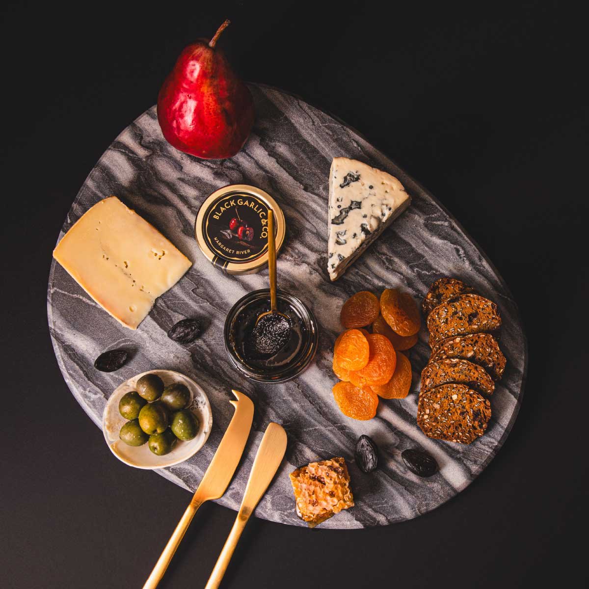 A stylised photograph of a marble cheese board with cheese, olives, crackers, dried apricots and Black Garlic & Co's savoury paste