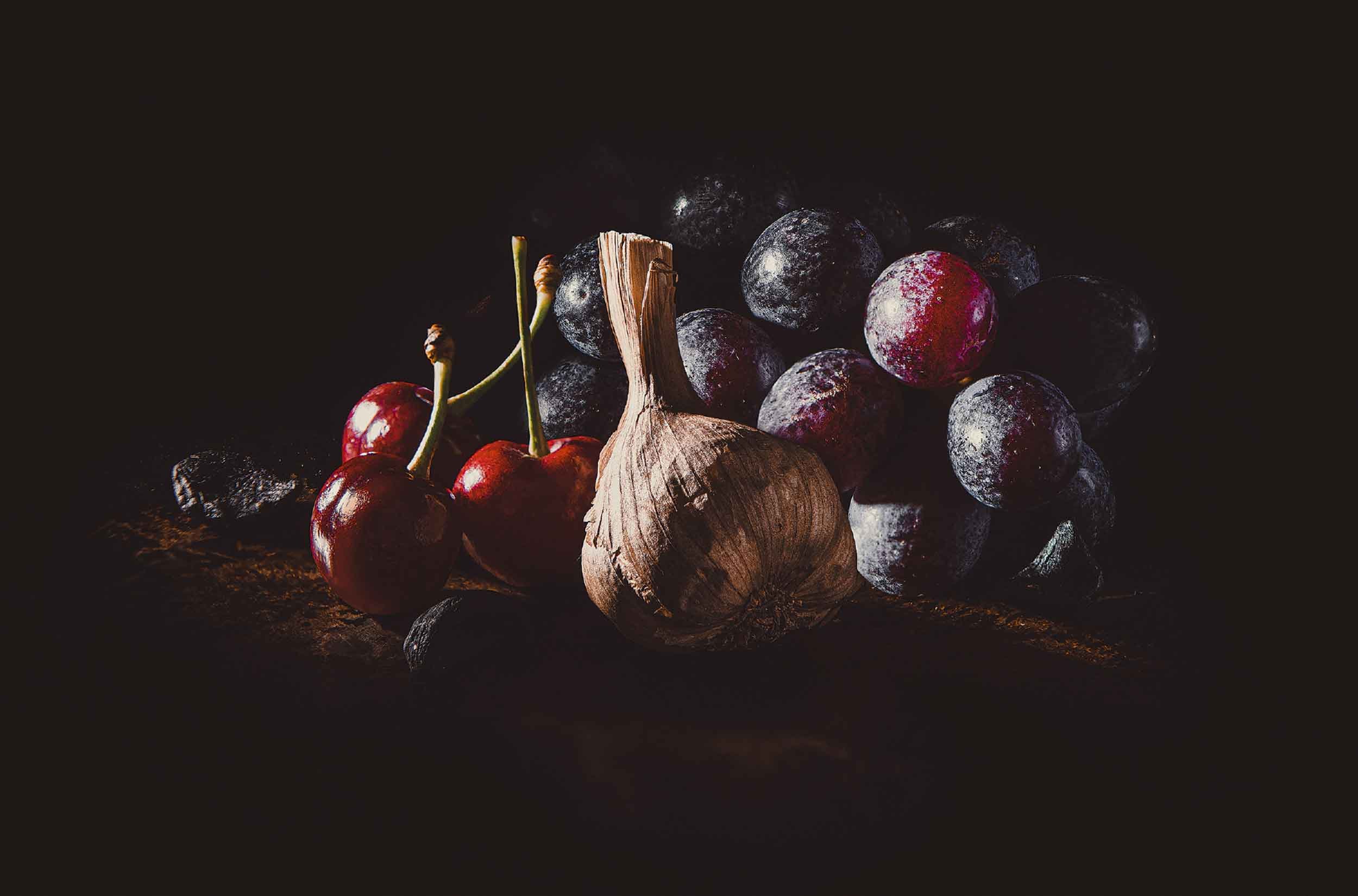 A stylised photograph for Black Garlic & Co, showcasing a black garlic bulb surrounded by cherries and red grapes