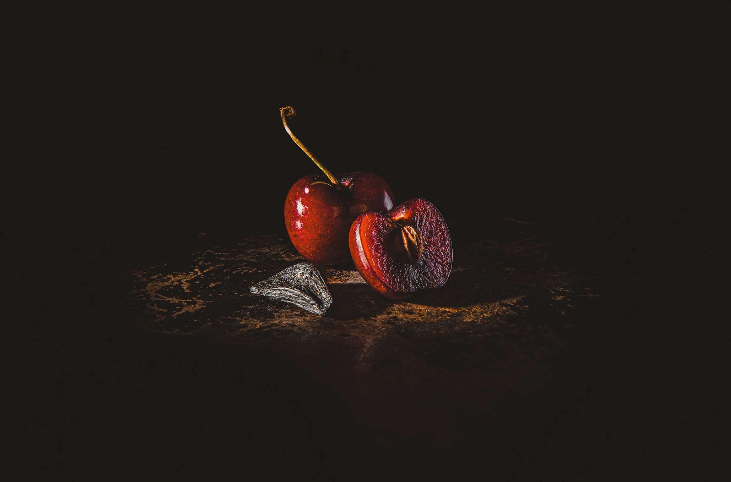 A stylised photograph for Black Garlic & Co showcasing a black garlic clove and two cherries, one that has been cut in half