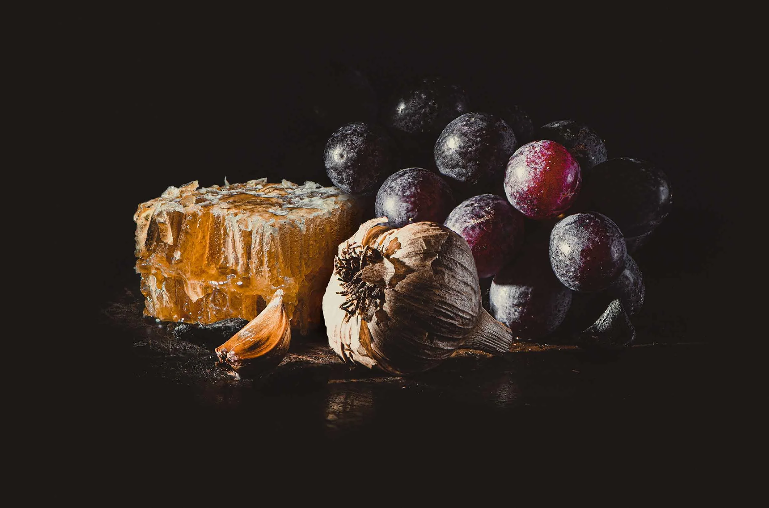 A stylised photograph for Black Garlic & Co showcasing honeycomb, black garlic and red grapes