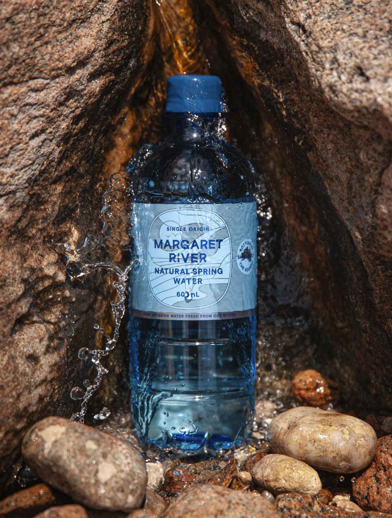 A 600ml Margaret River Natural Spring Water bottle sitting on a rock bed with water splashing over the top of it