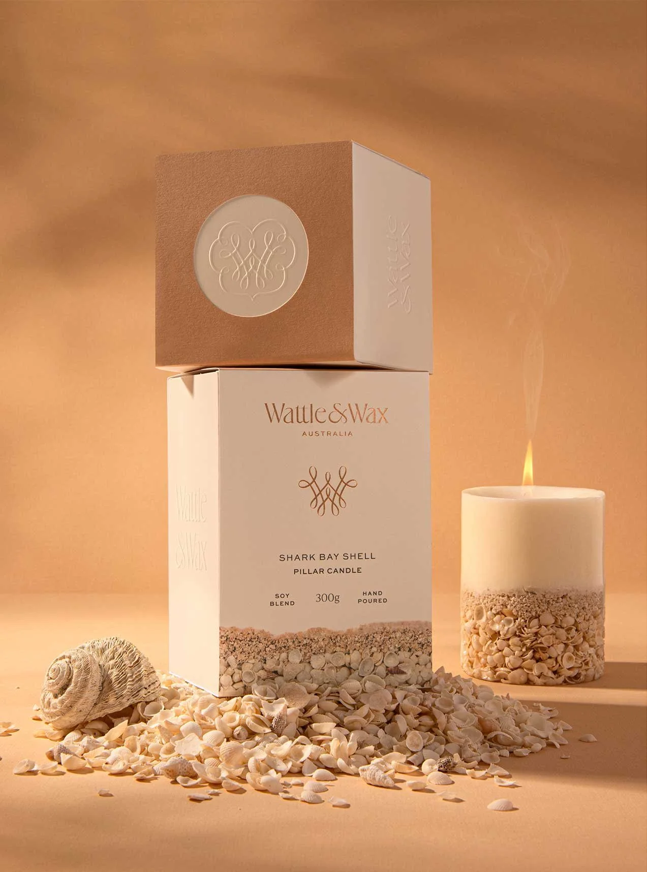 A photograph of Wattle and Wax's Shark Bay Pillar Candle packaging in the foreground with a candle burning to the side