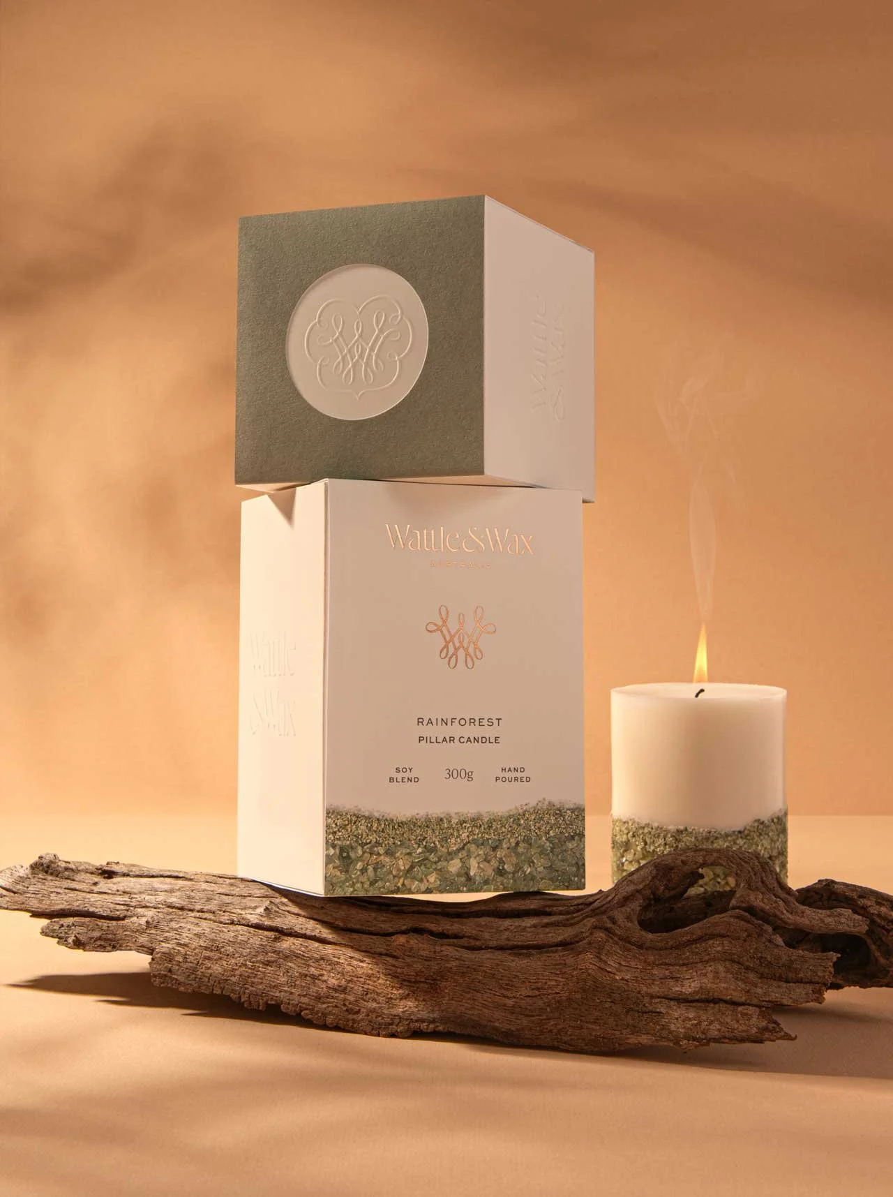 A stylised photograph of Wattle and Wax Australia's Rainforest pillar candle, with two candle packaging boxes sitting on top of a piece of drift wood and a candle burning
