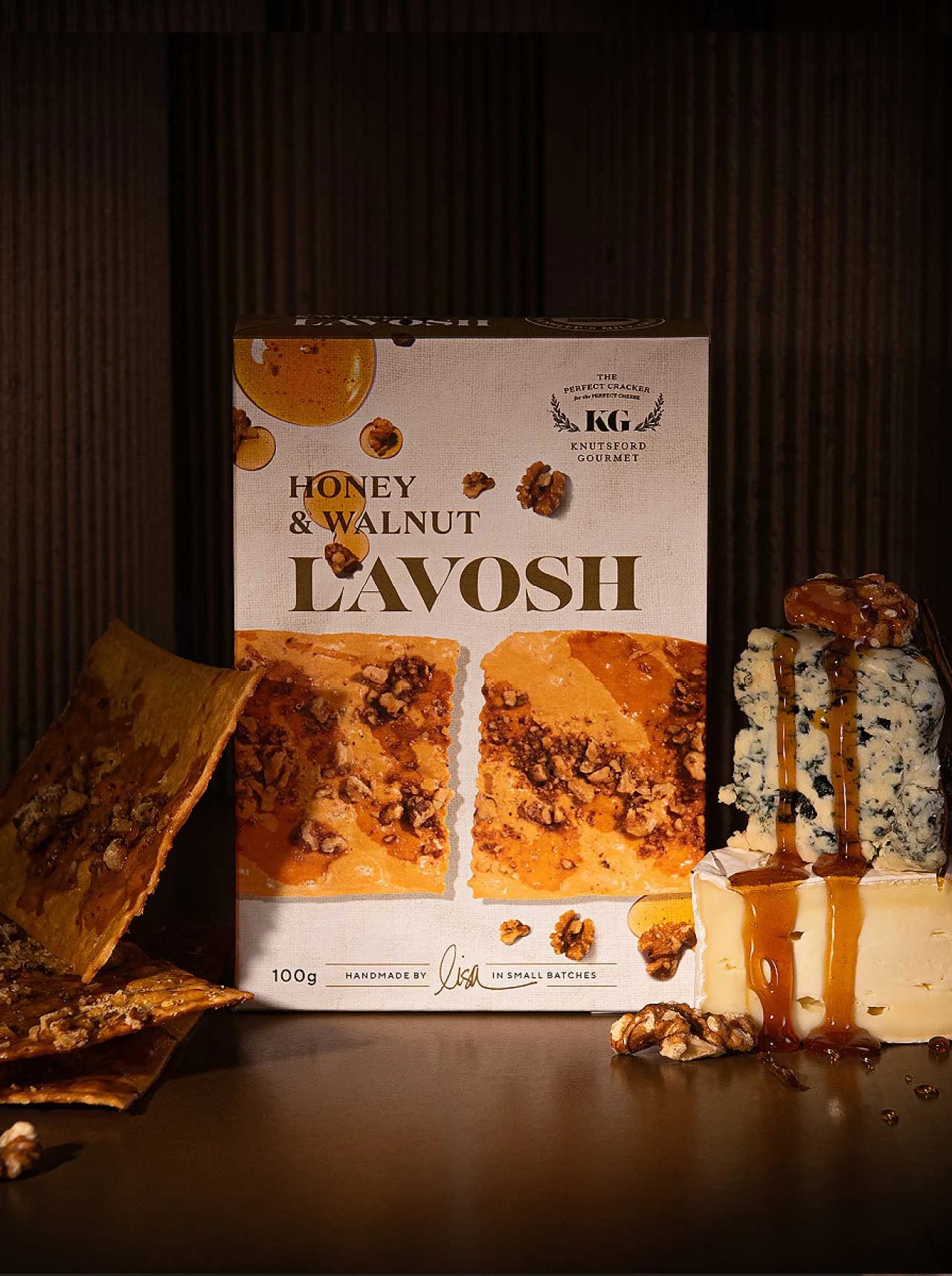 A stylished photograph of Knutsford Gourmet's Honey and Walnut Lavoush packaging, surrounded by blue cheese and brie, with honey drizzling over the side of the cheeses and the lavoush positioned next to the packaging box