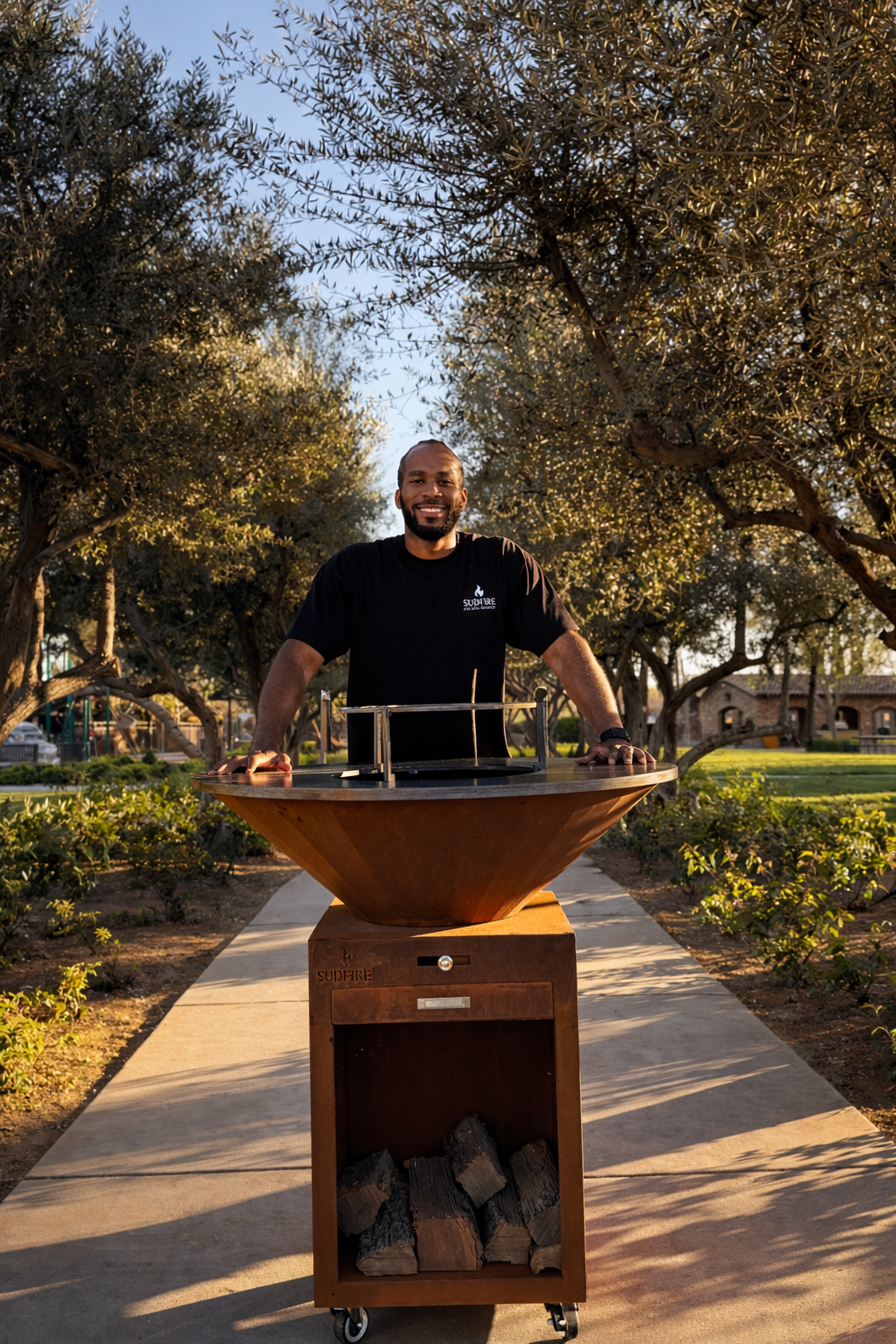 Man standing outdoors behind a wood-fired pizza oven in a park with trees and buildings in the background at sunset.