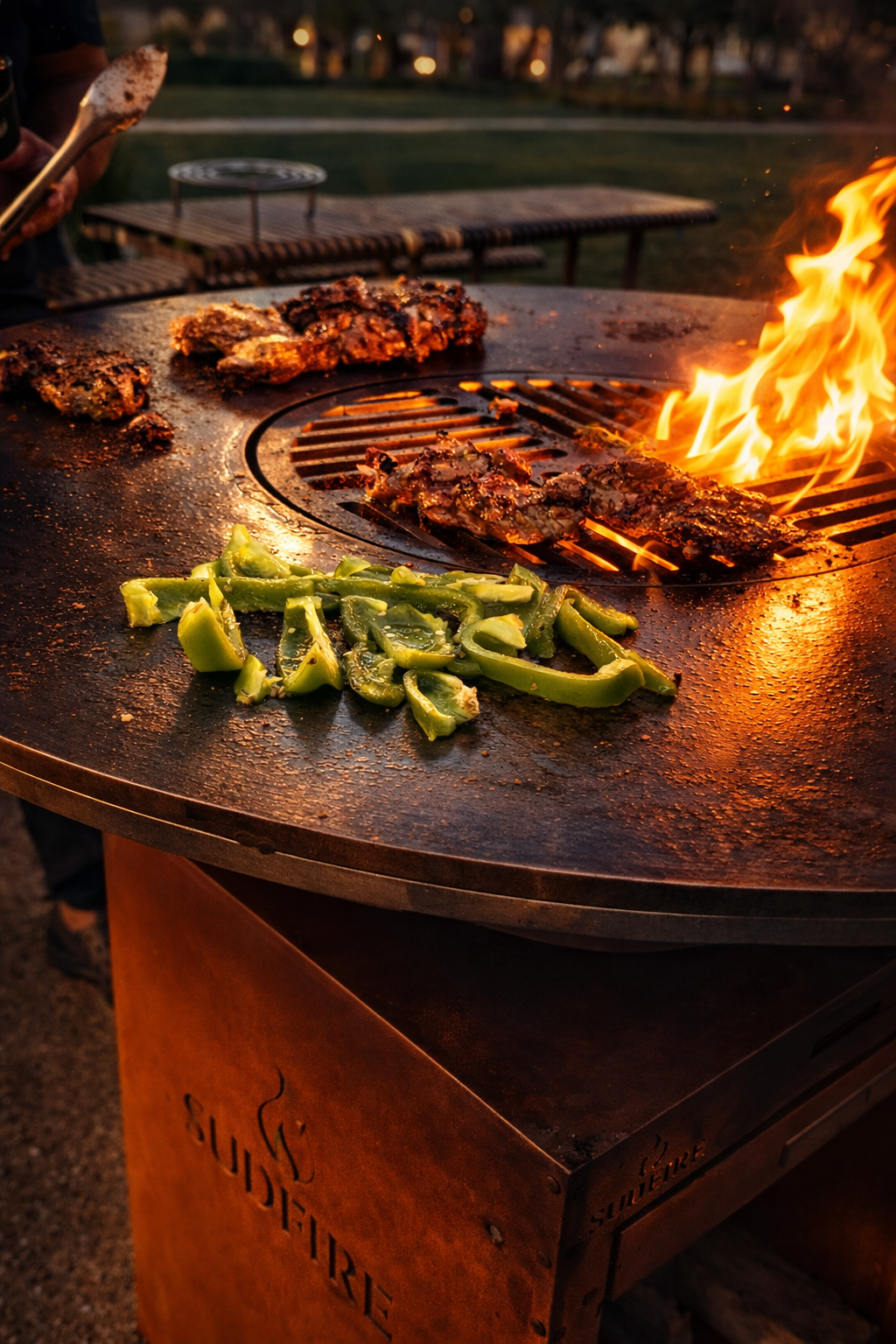 Barbecue grill with flames, cooked meats, and green bell peppers on a rustic outdoor barbecue at sunset.