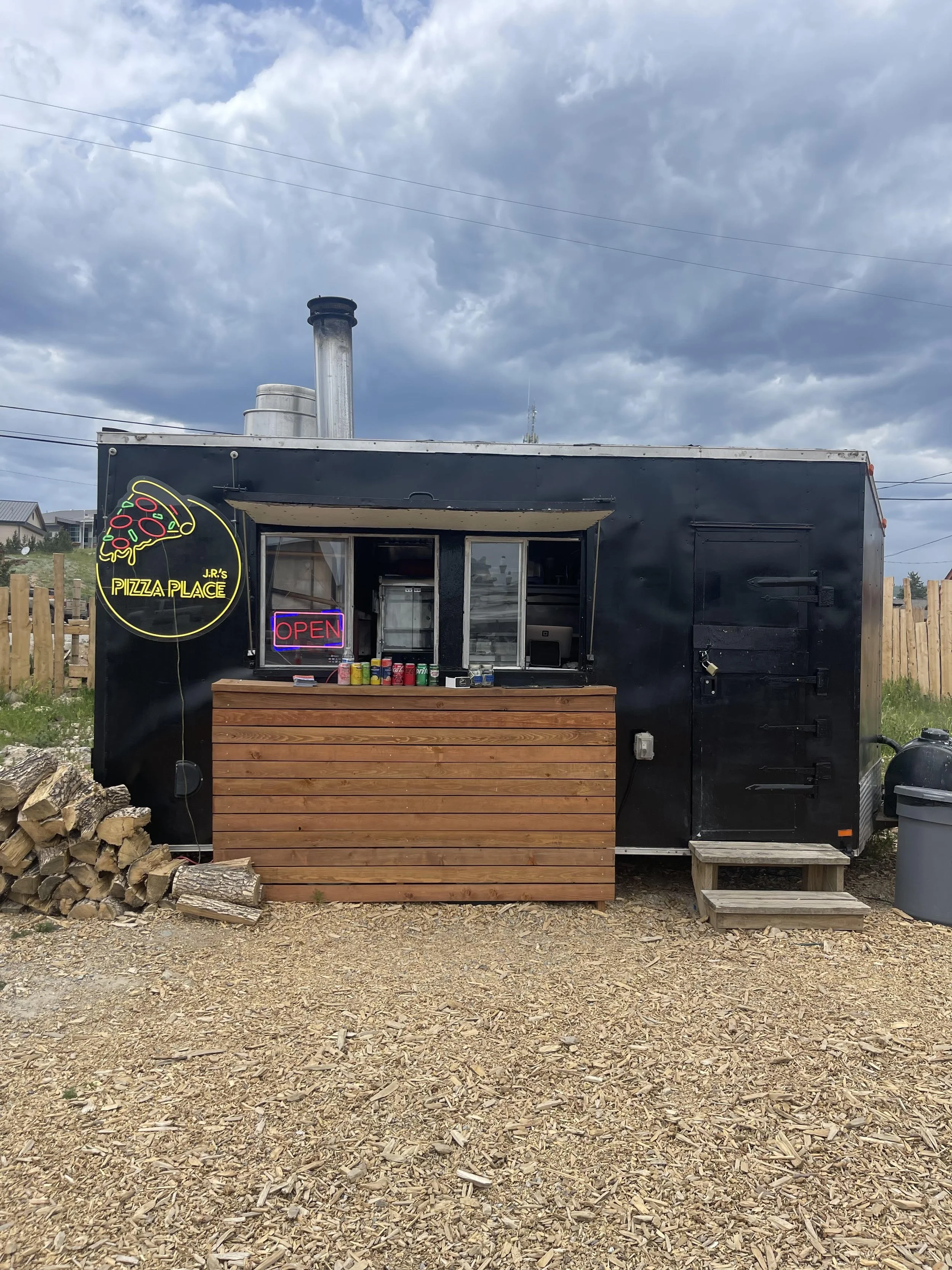 Food truck named 'J.R.'s Pizza Place' with a neon 'Open' sign, serving pizzas, parked on a gravel area under cloudy sky, with firewood stacked nearby.