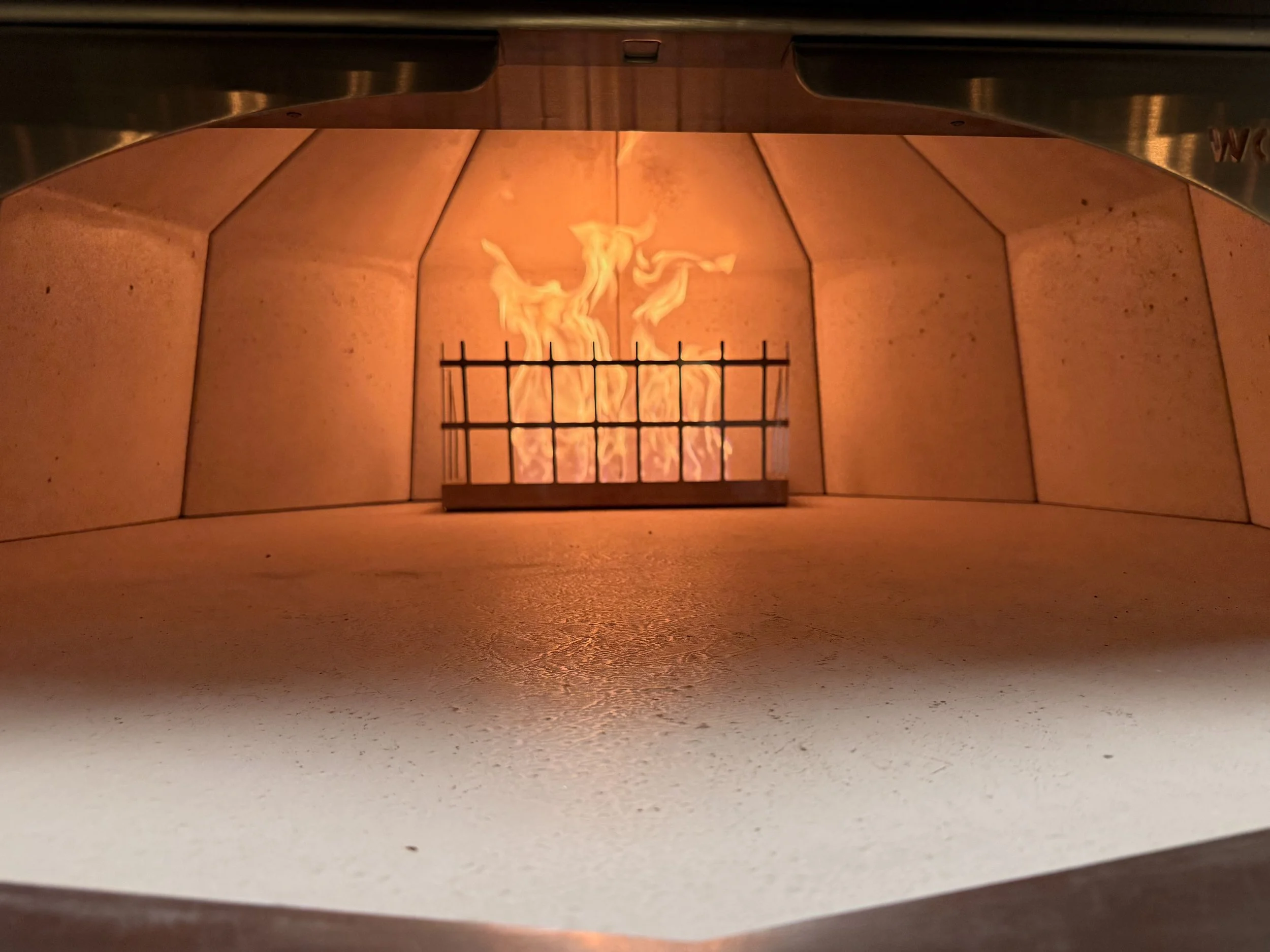 Inside a pizza oven with a visible fire burning behind a metal grill, surrounded by pinkish tiles.