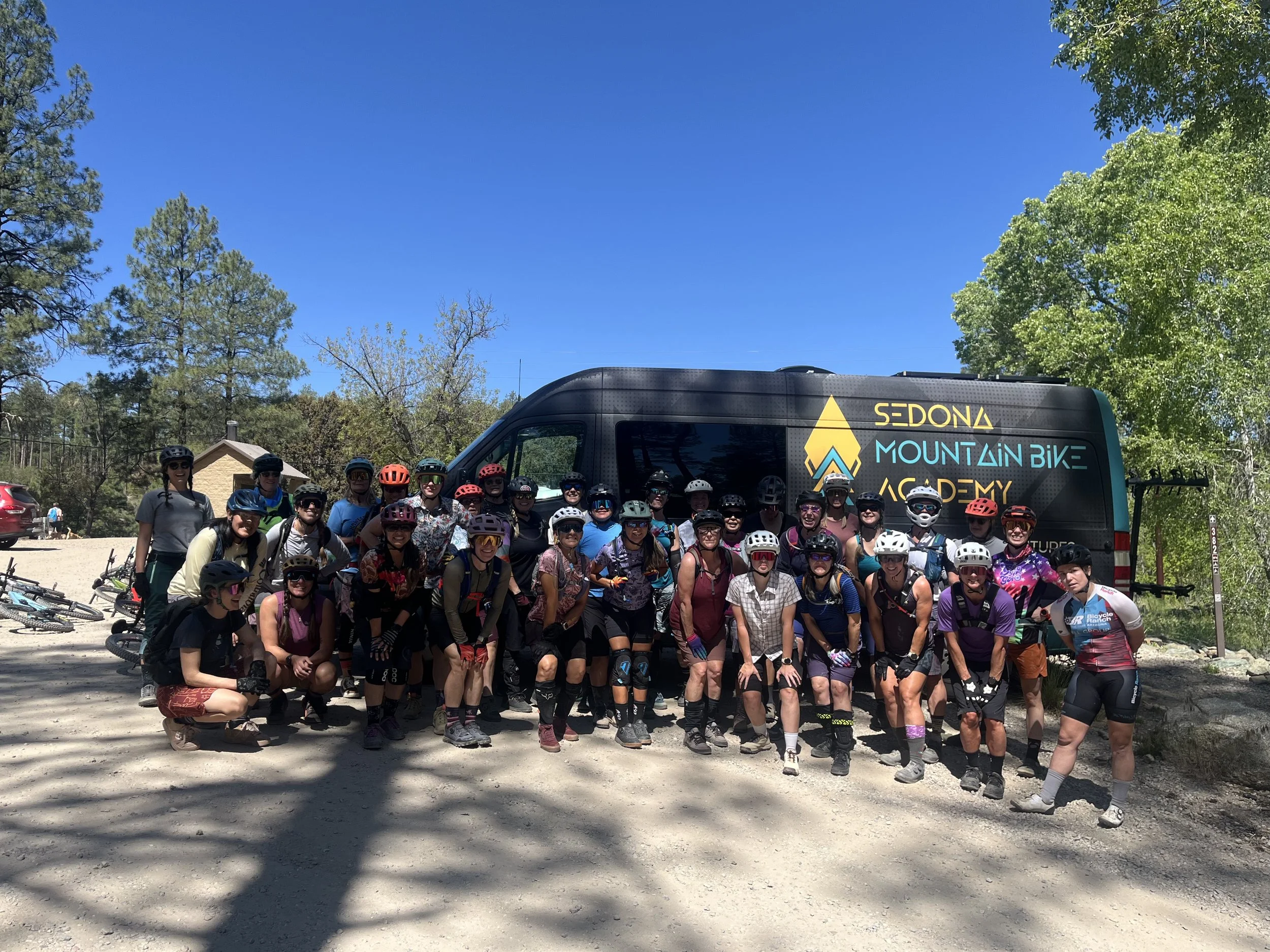 Group of people in cycling gear posing outdoors in front of a large van with a sign that says Sedona Mountain Bike Academy, surrounded by trees and clear blue sky.